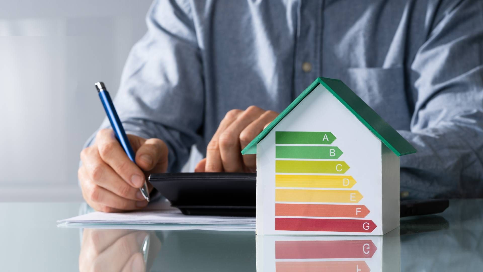 Person calculating with a pen and calculator near a house model with an energy efficiency rating.