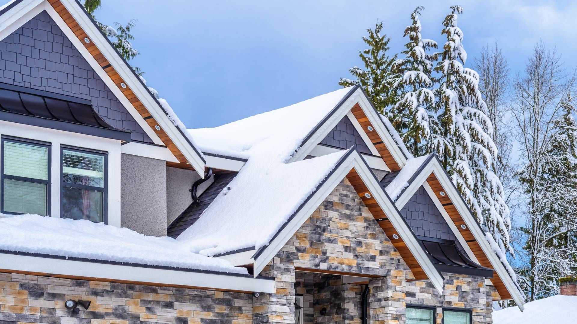 Snow-covered house with stone facade, dark roof, and wooden accents, set against snowy trees and a blue sky.