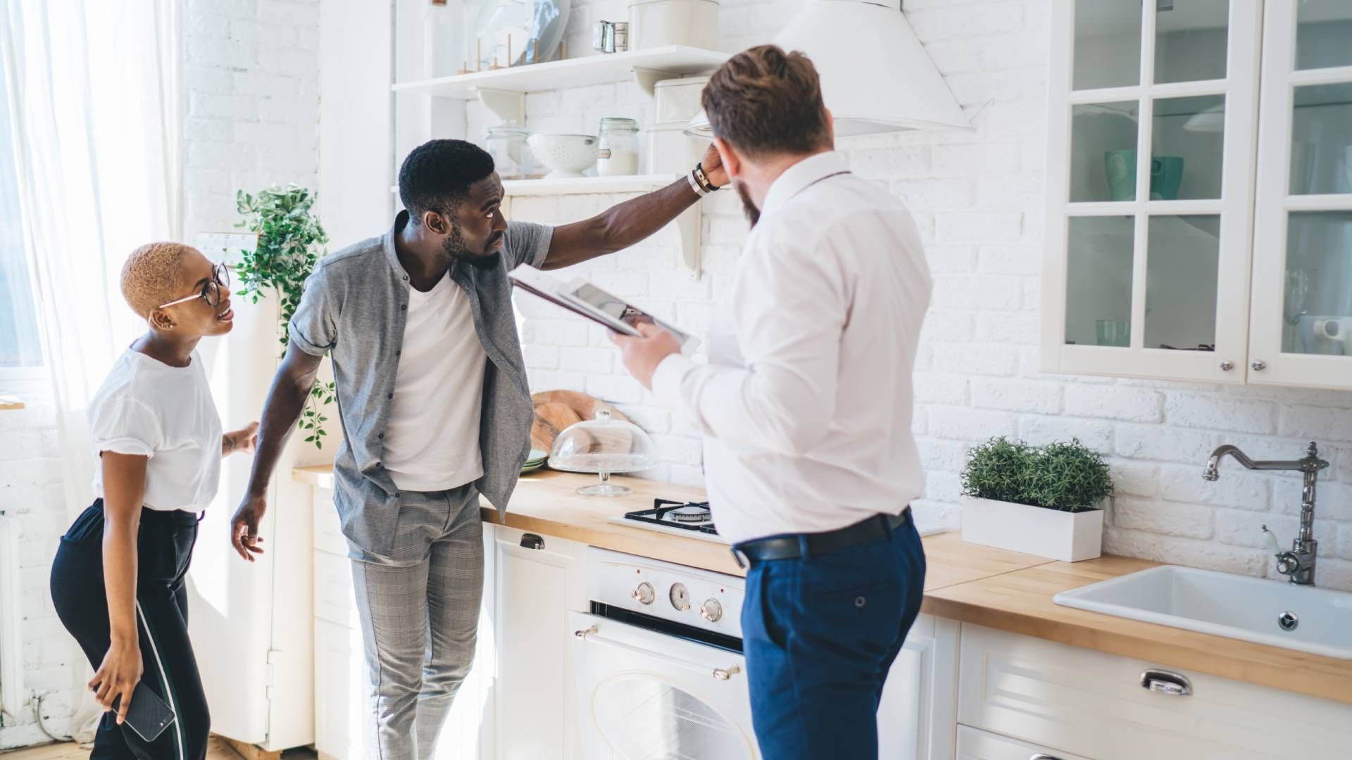 Couple and realtor in a bright kitchen; man points at hood, woman looks on.