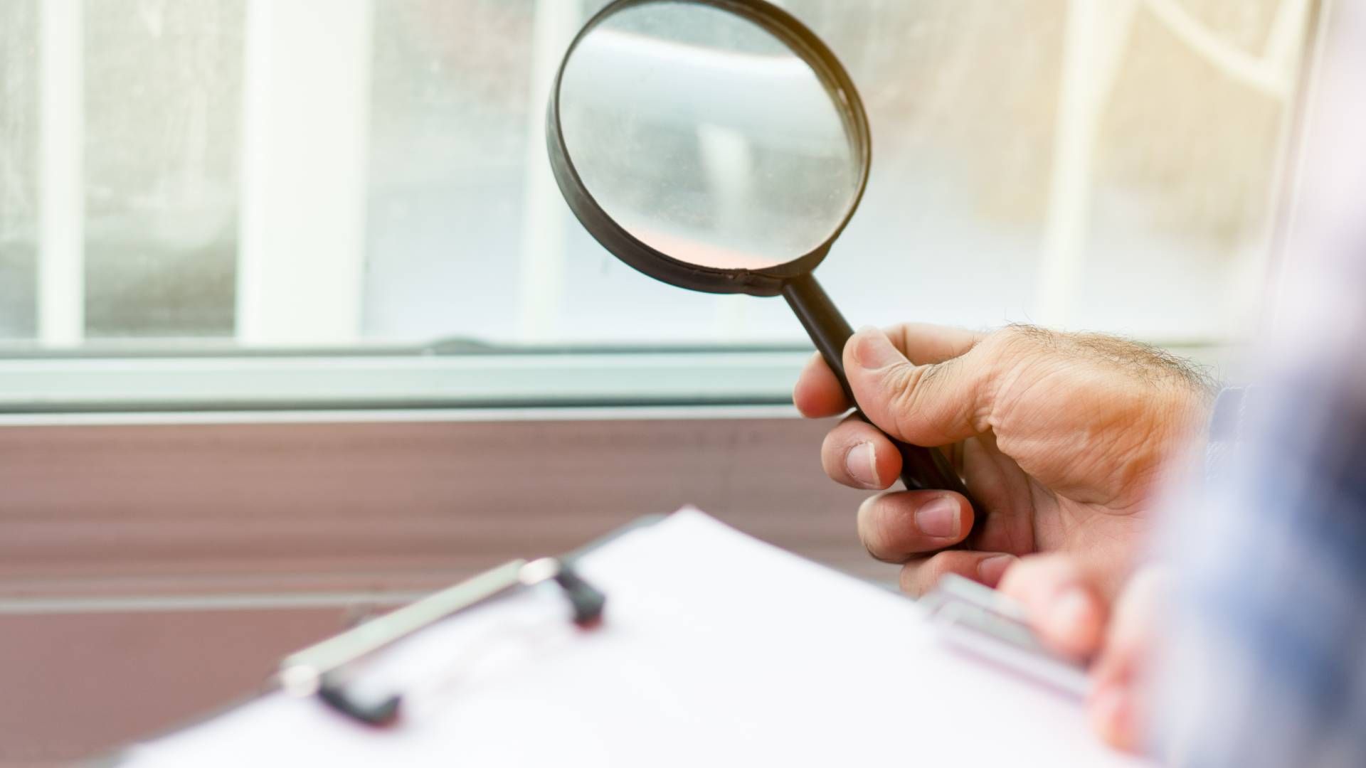 Hand holding magnifying glass, inspecting window, clipboard in foreground.