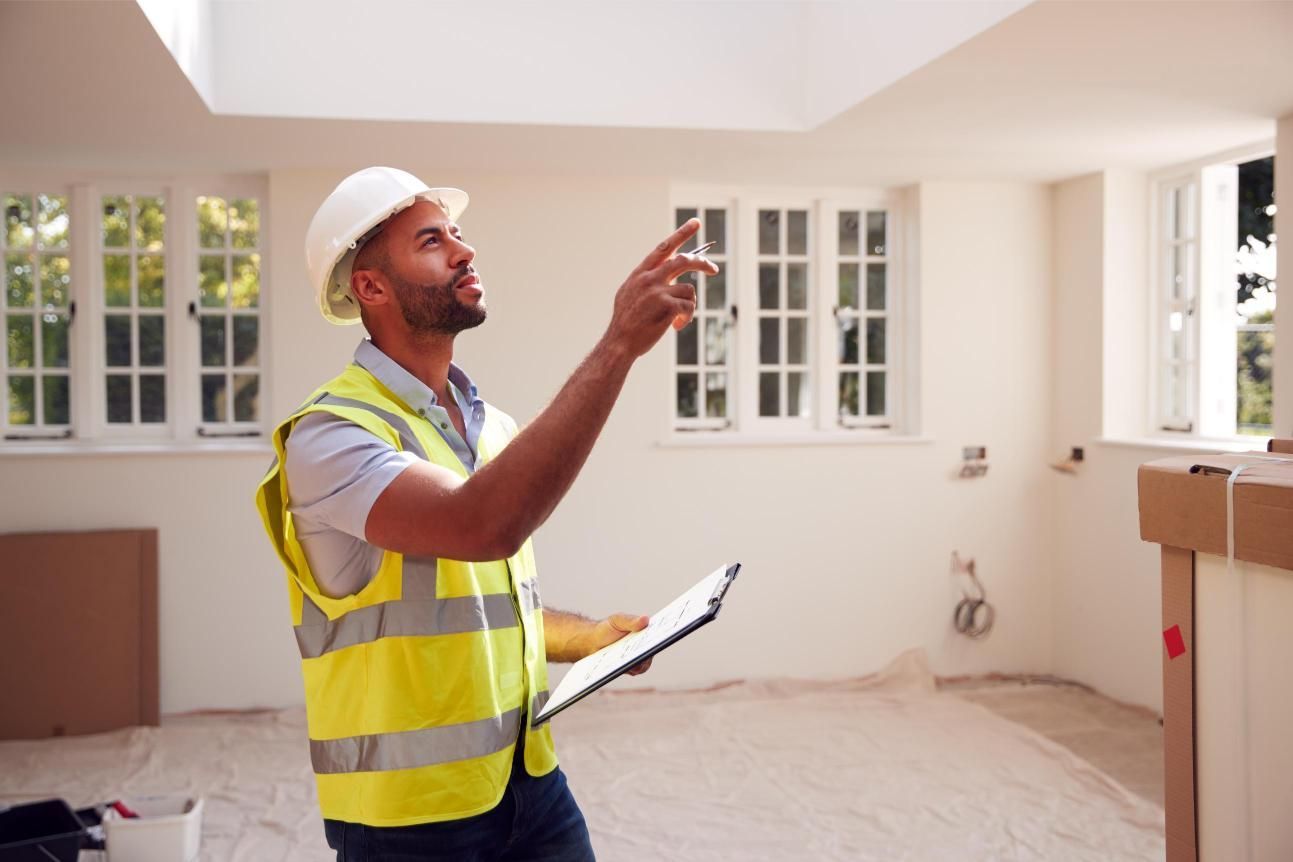 Construction worker in a safety vest and helmet inspecting a room, pointing upward with a clipboard.