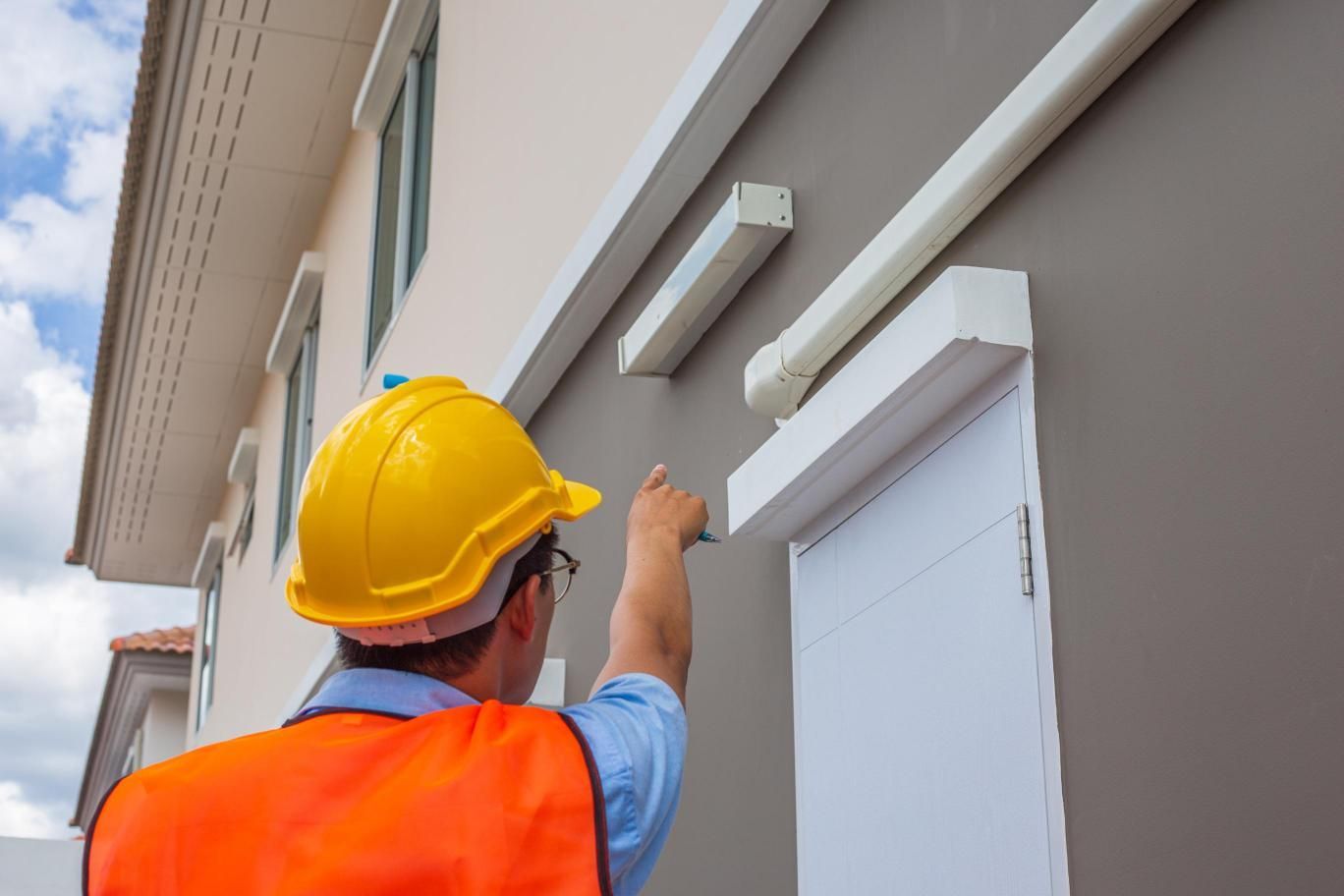 Person in safety gear inspecting a house exterior, pointing at the wall.