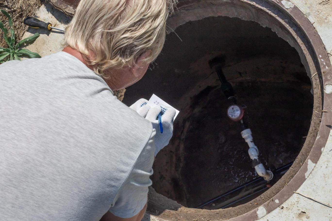 Person in gray shirt looks into open manhole, holding papers.