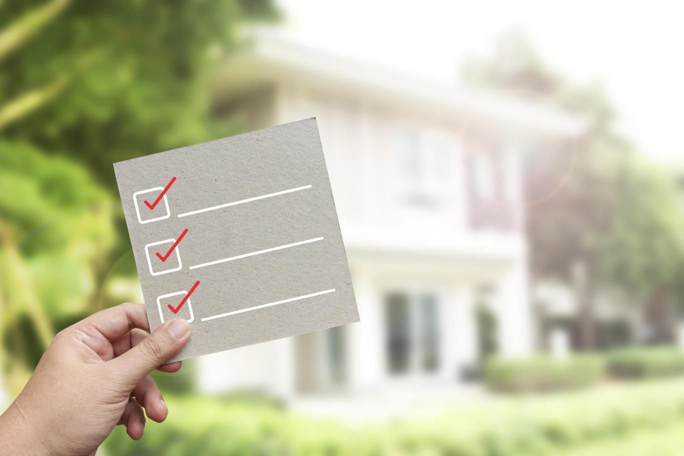 Hand holding a checklist with red checkmarks in front of a blurred house.
