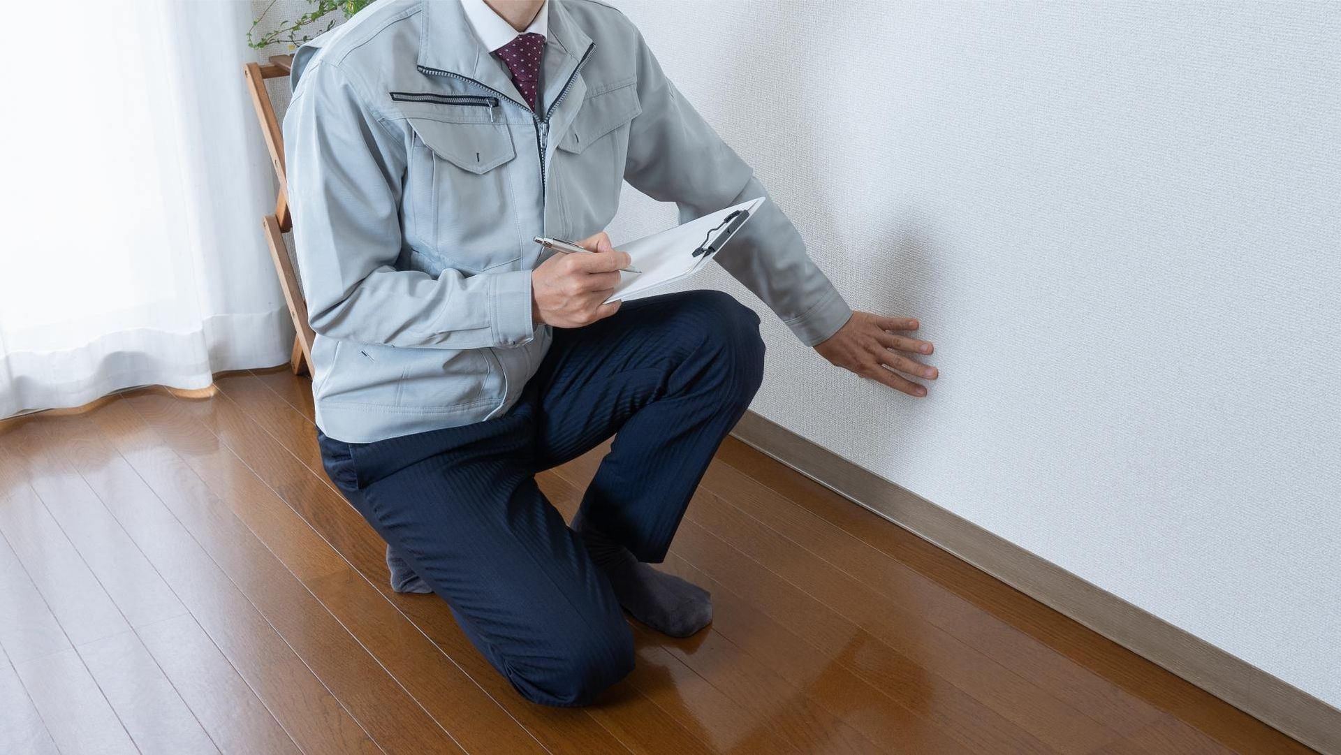 Person in work attire inspecting a wall, holding a clipboard, in a room with wood flooring.