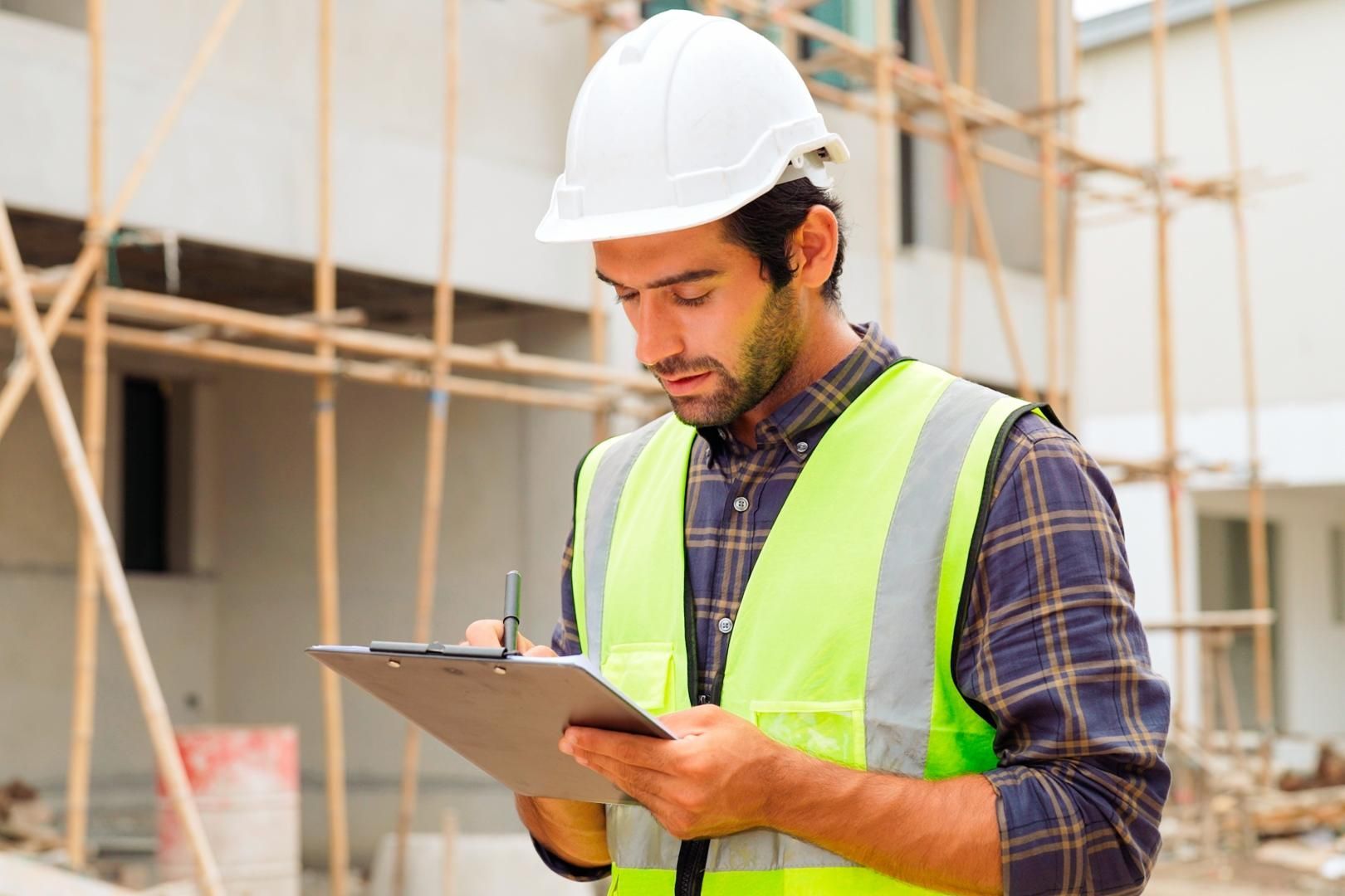 Construction worker in hard hat and vest taking notes on a clipboard at a construction site.