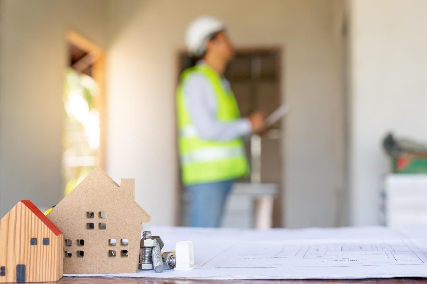Miniature wooden houses on blueprints; construction worker inspecting a building in background.