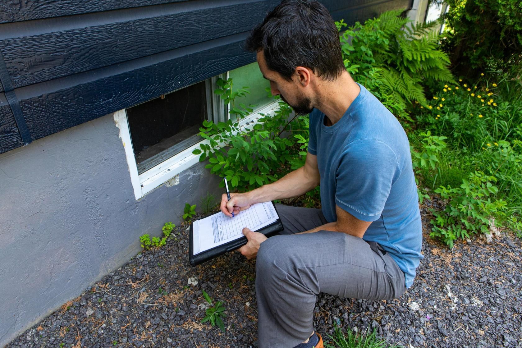 Man crouches beside a building, writing on a clipboard. A window is visible.