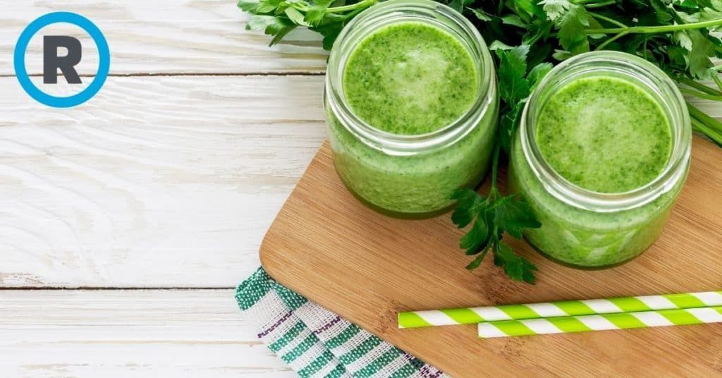 Two jars of green smoothie with straws on a wooden cutting board.