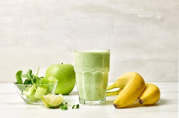 A green smoothie in a glass with bananas , apples and mint on a table.