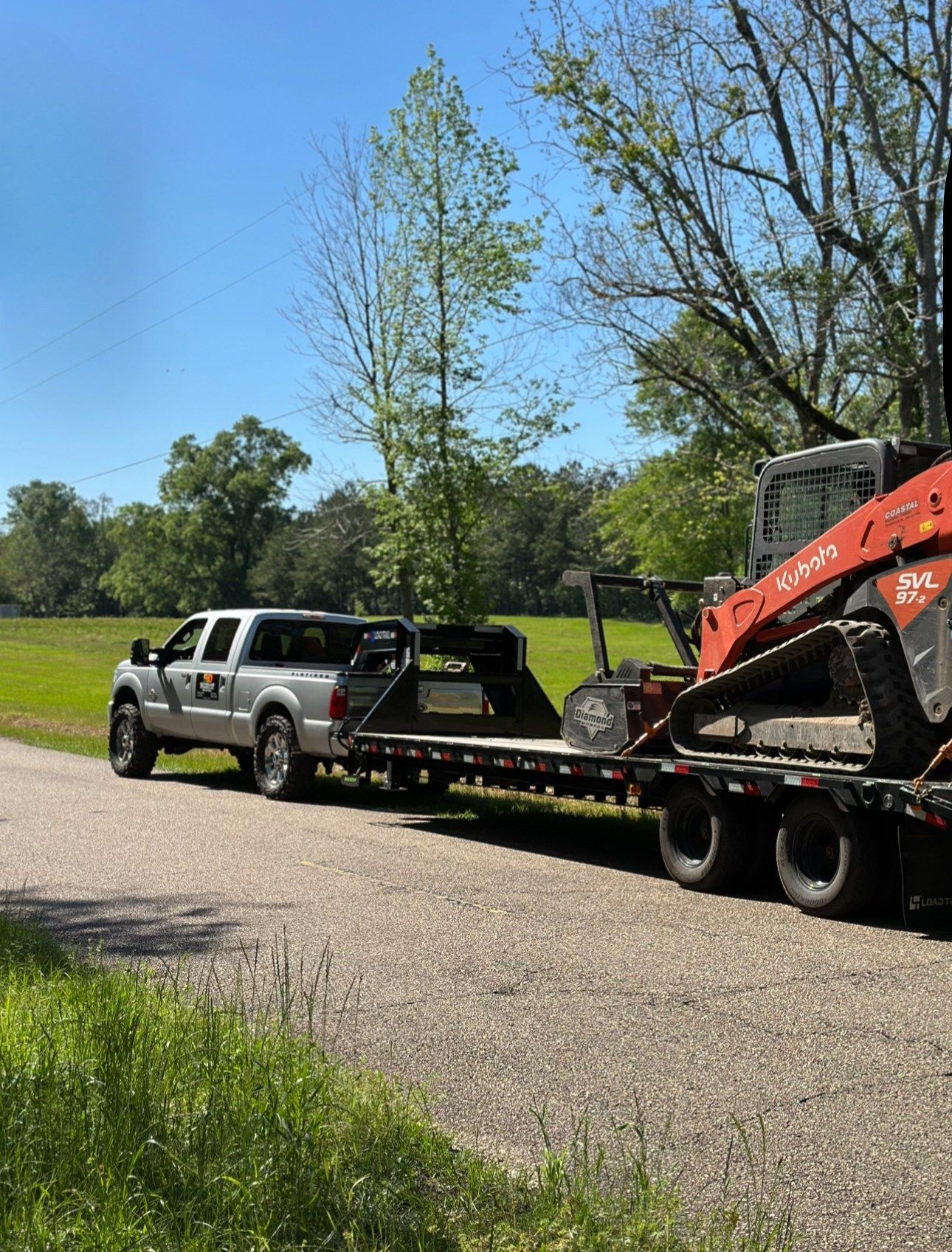 A silver pickup truck towing a flatbed trailer with a red Kubota skid-steer loader on a gravel path near trees.