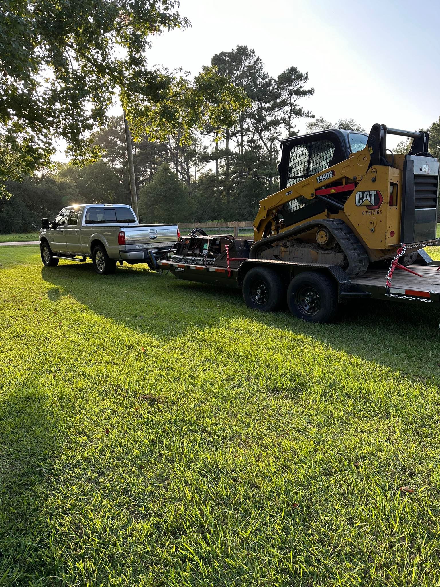 A tan pickup truck pulls a trailer carrying a yellow Caterpillar tracked skid steer across a grassy field.