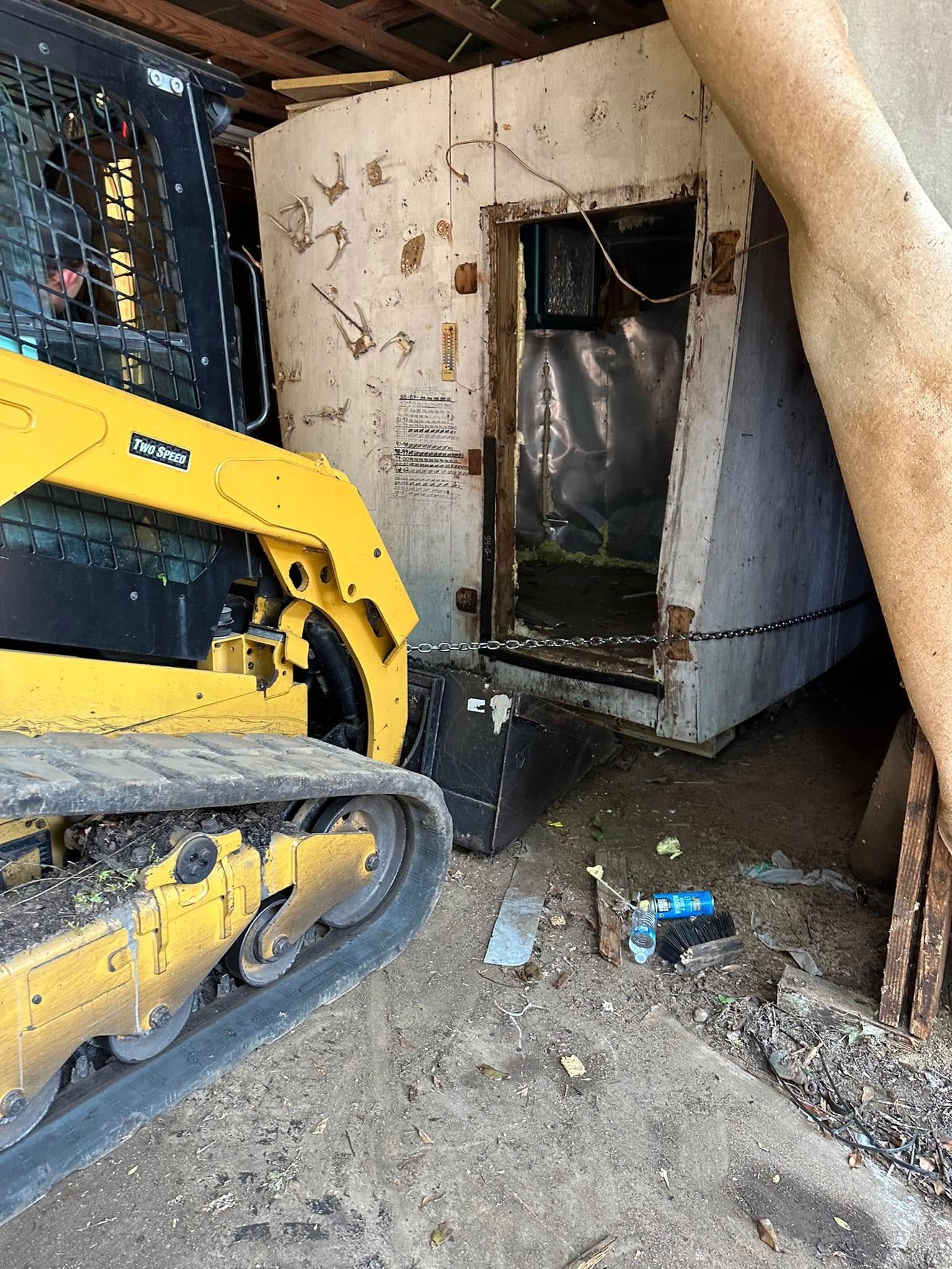 A yellow skid steer loader sits near a partially demolished white structure, with debris on the ground.