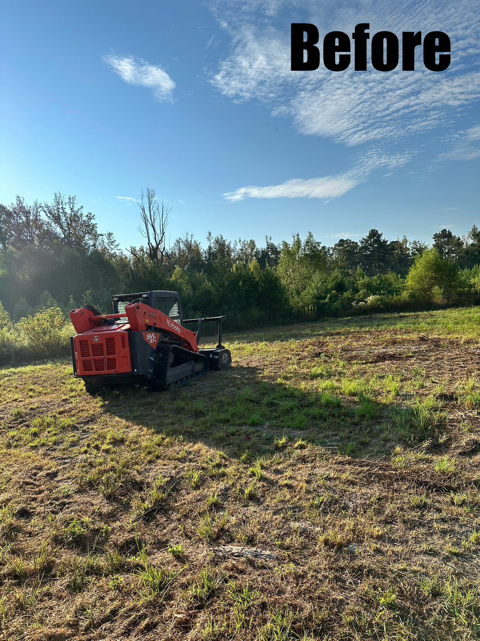 An orange skid steer with a forestry mulcher attachment working in a field with a treeline under a sunny sky.