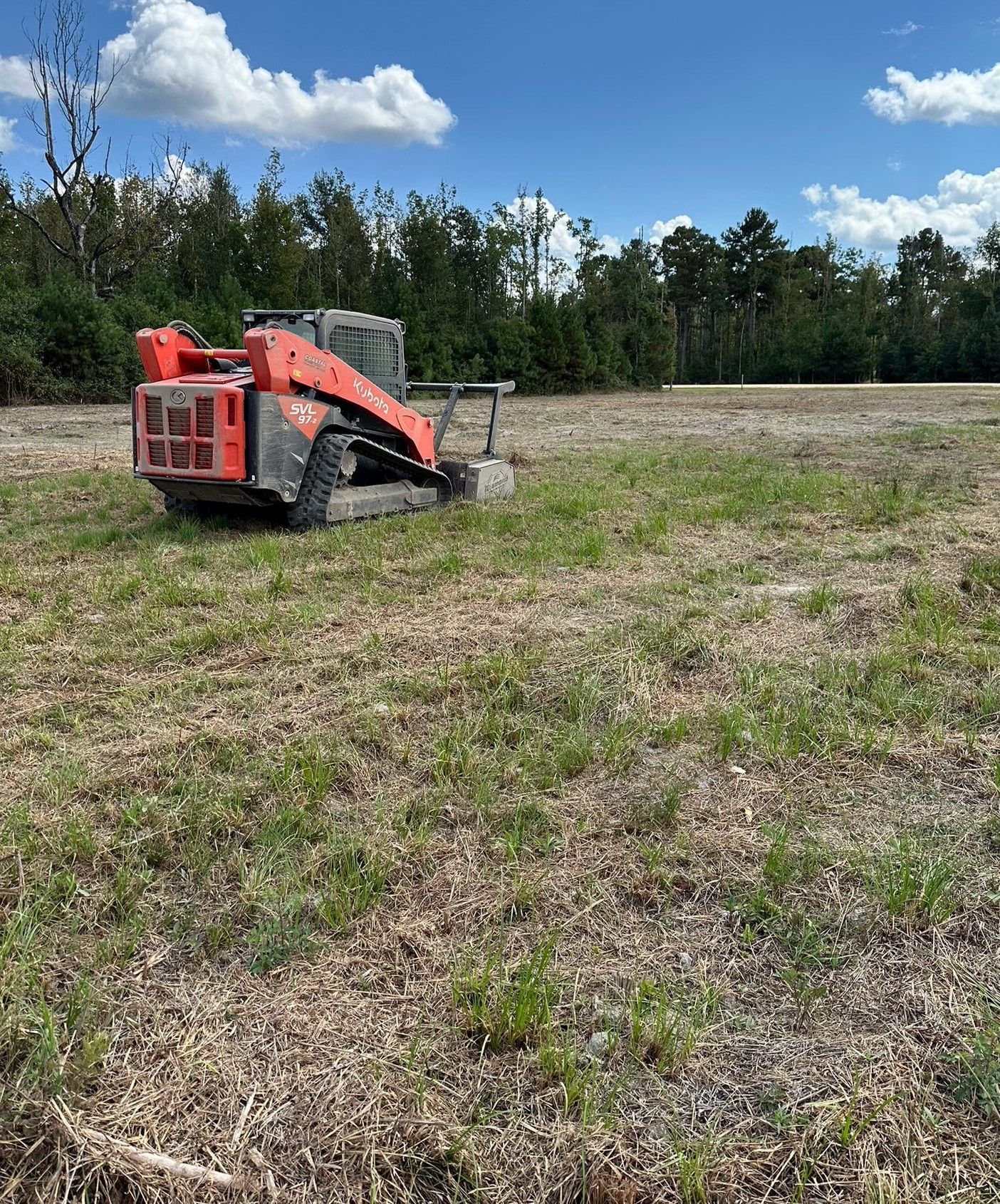 An orange tracked skid steer sits on a large, flat, cleared field under a bright blue sky with scattered clouds.