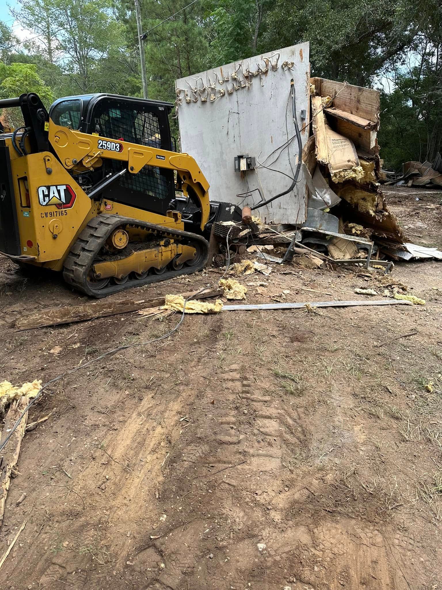 A yellow Caterpillar skid steer loader pushes a large section of white, damaged wall in a wooded area.