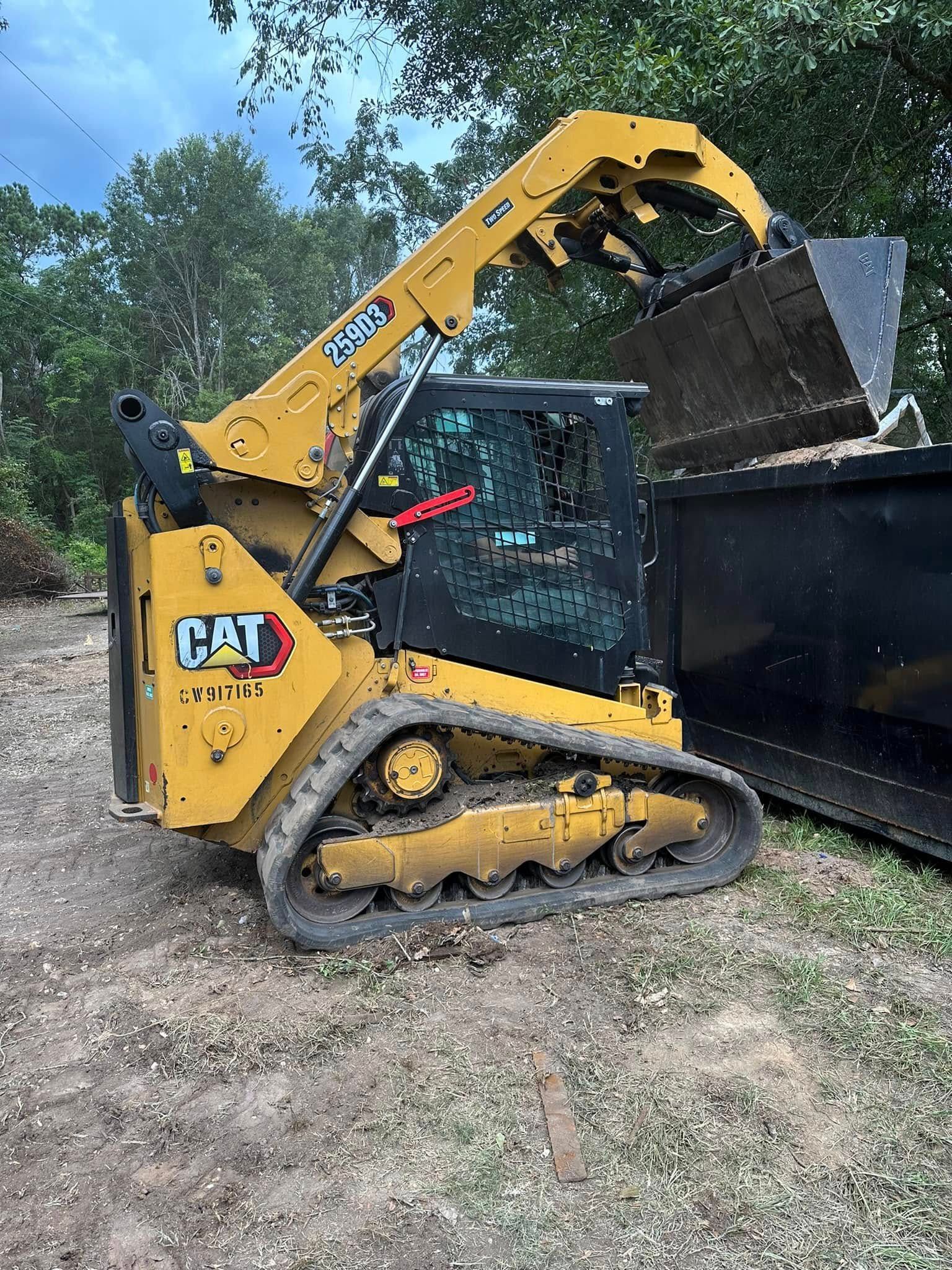 A yellow Caterpillar compact track loader dumping material into a large black metal container on a dirt site.