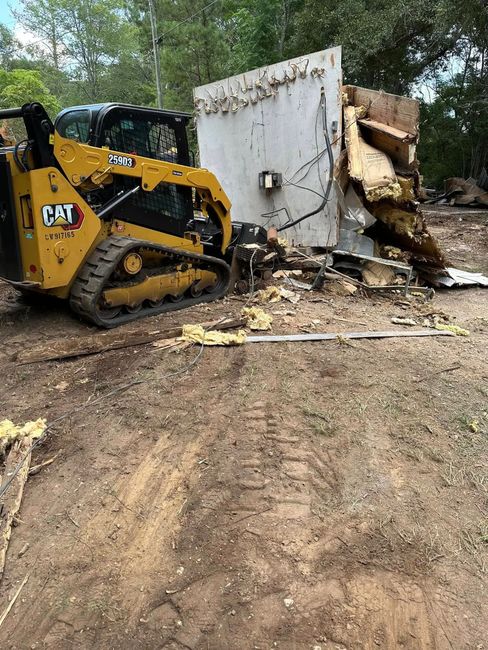 A yellow CAT compact track loader pushes a section of a white, partially demolished wall in a wooded area.