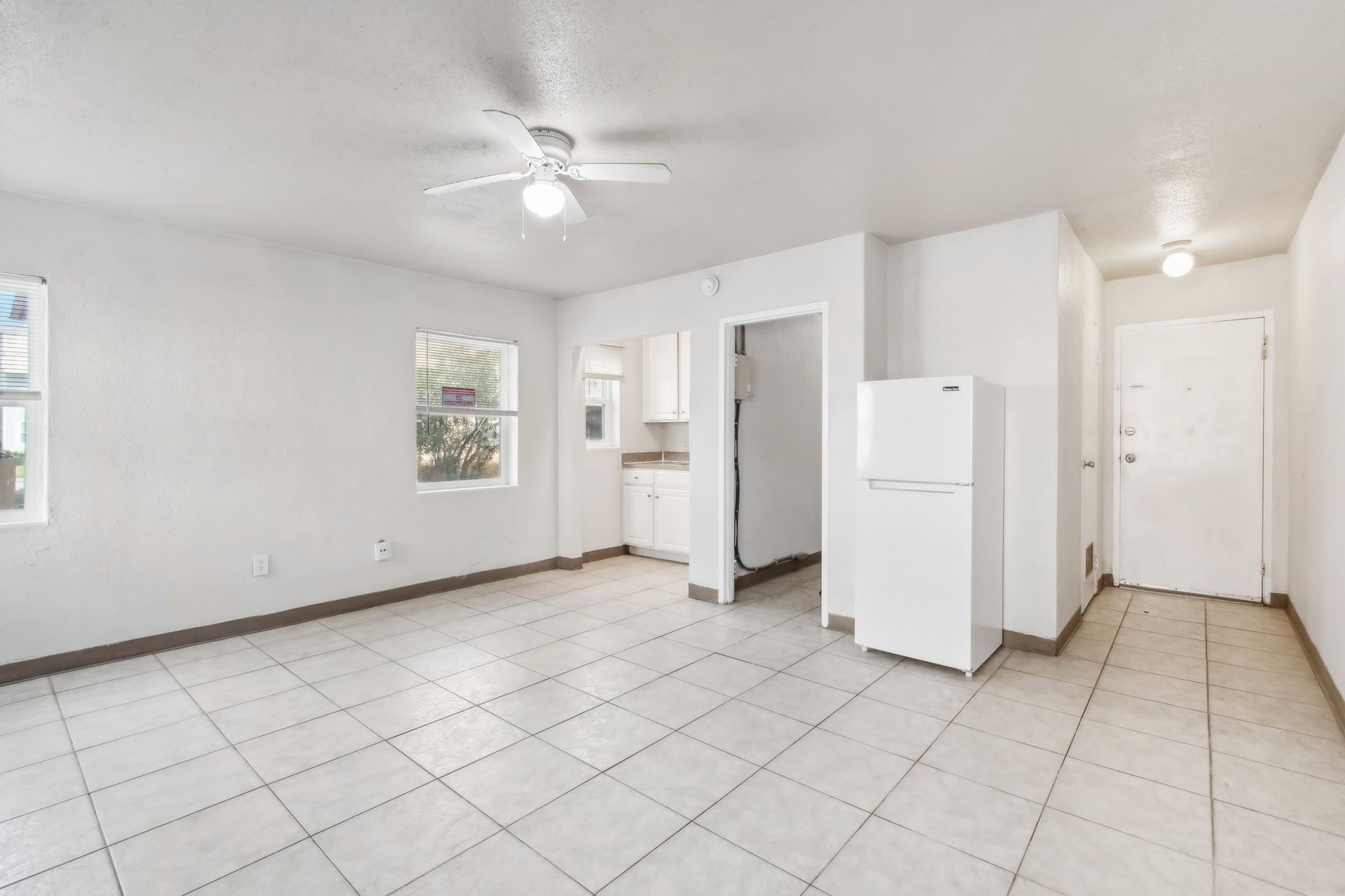 White-walled apartment interior with tile floor, two windows, refrigerator, kitchen doorway, and ceiling fan.