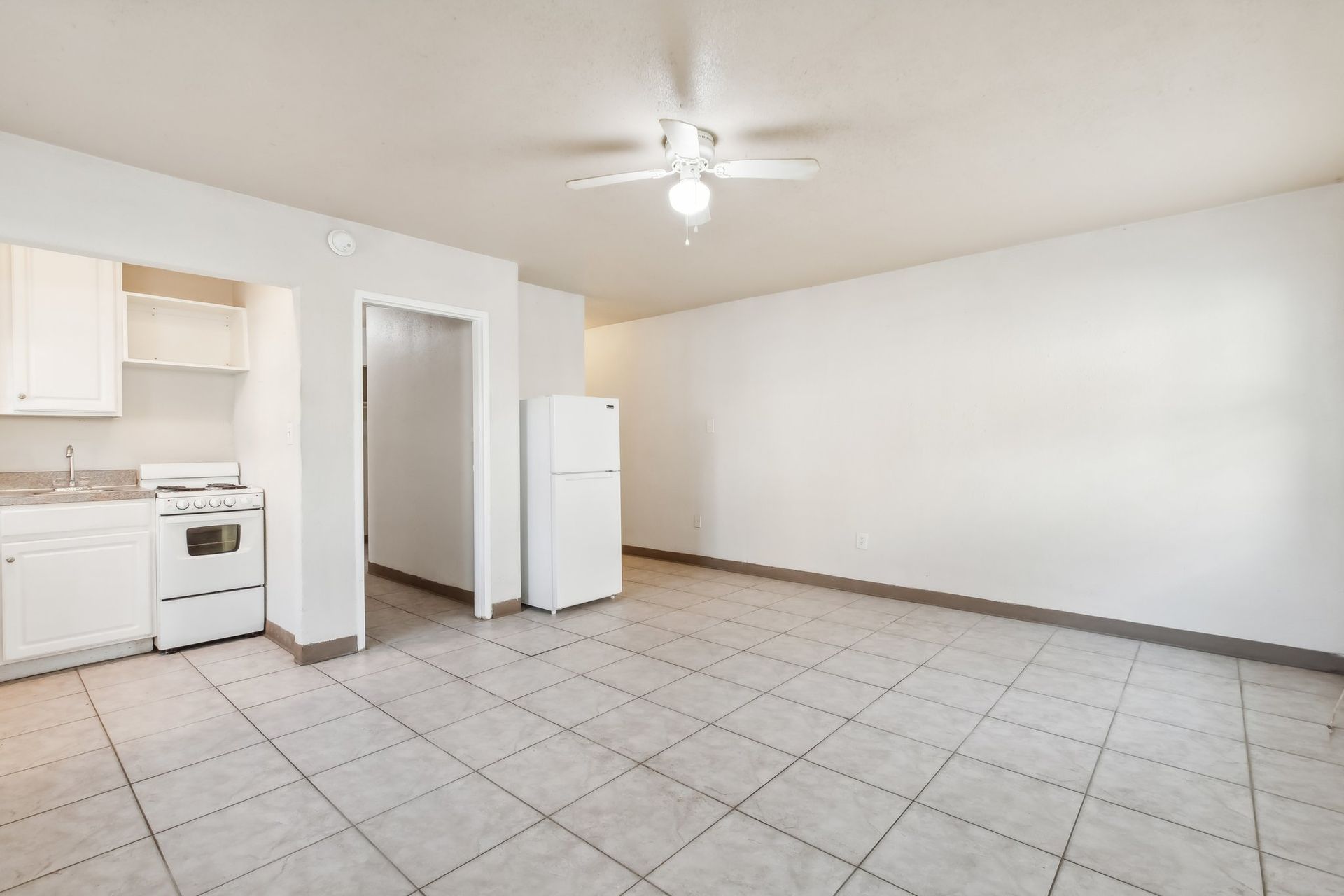 Empty apartment interior with kitchen and living space, white walls, tile floor, and refrigerator.