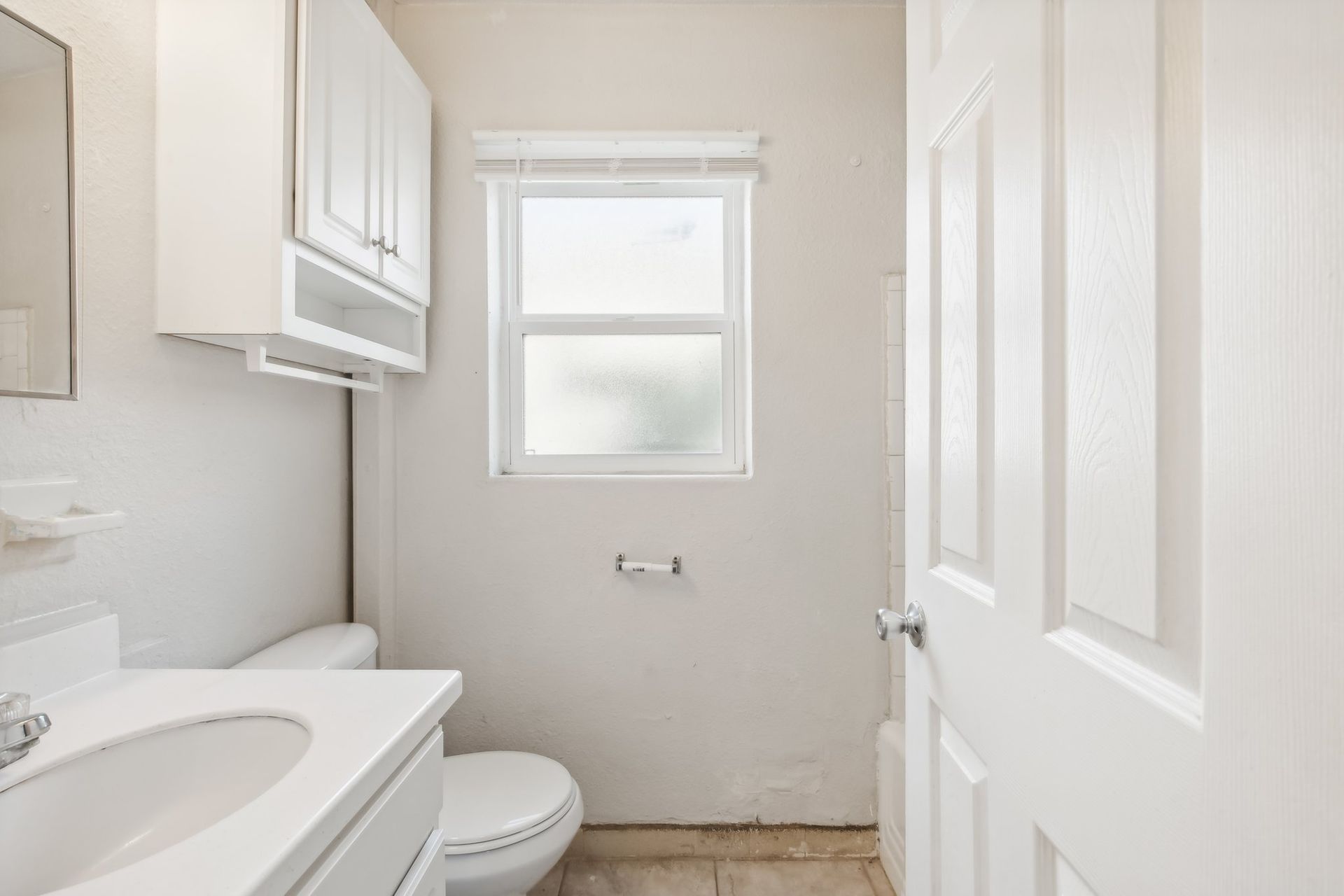 Small, white bathroom with toilet, sink, and window. Overhead cabinet, light-colored walls, and closed door.