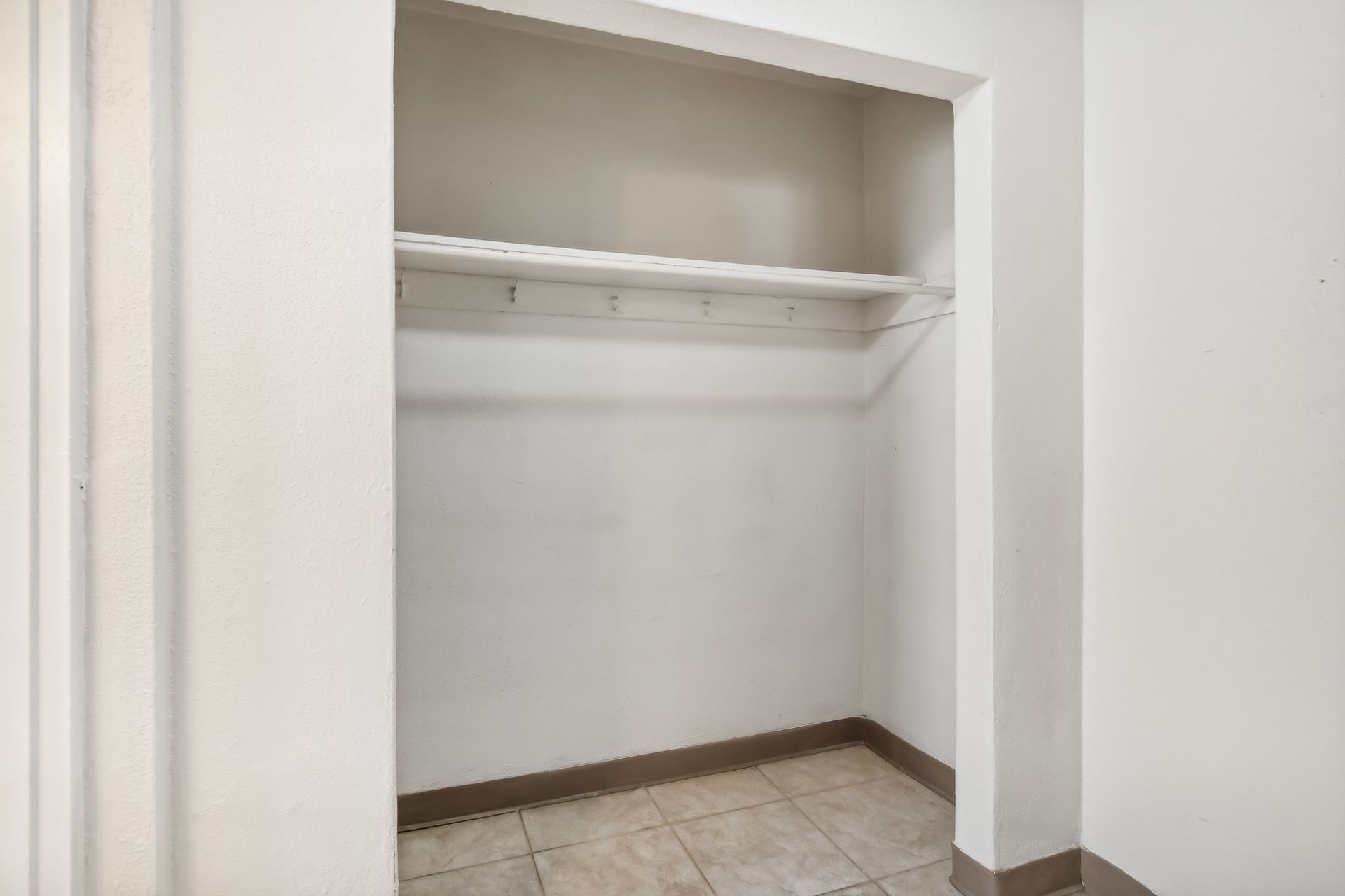 Empty white closet with a shelf, tile floor, and brown trim.
