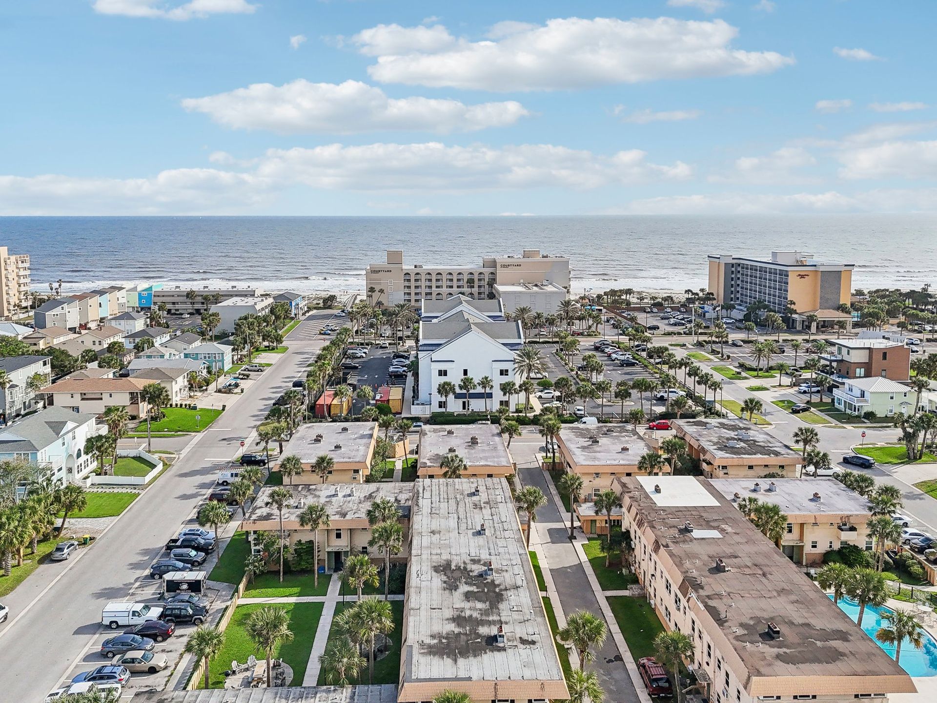 Aerial view of coastal town with beach, buildings, roads, and ocean under a partly cloudy sky.