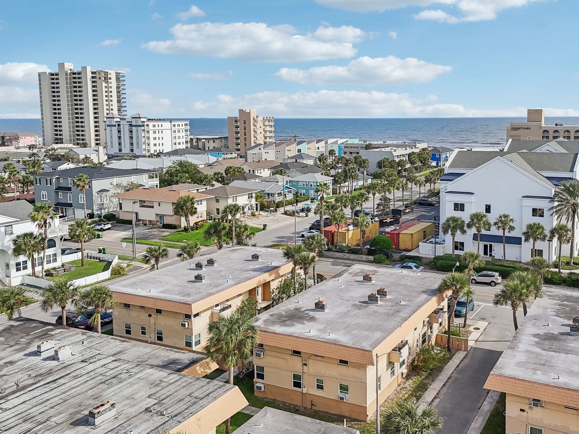 Aerial view of a coastal town with buildings, palm trees, and the ocean in the background under a blue sky.