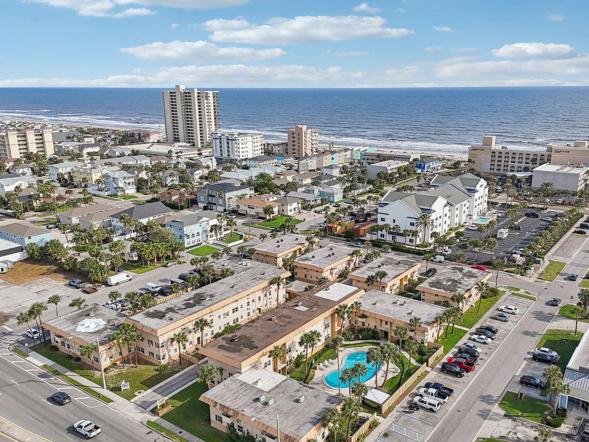 Aerial view of a coastal town with buildings, ocean, and a blue sky. Palm trees and a pool are visible.
