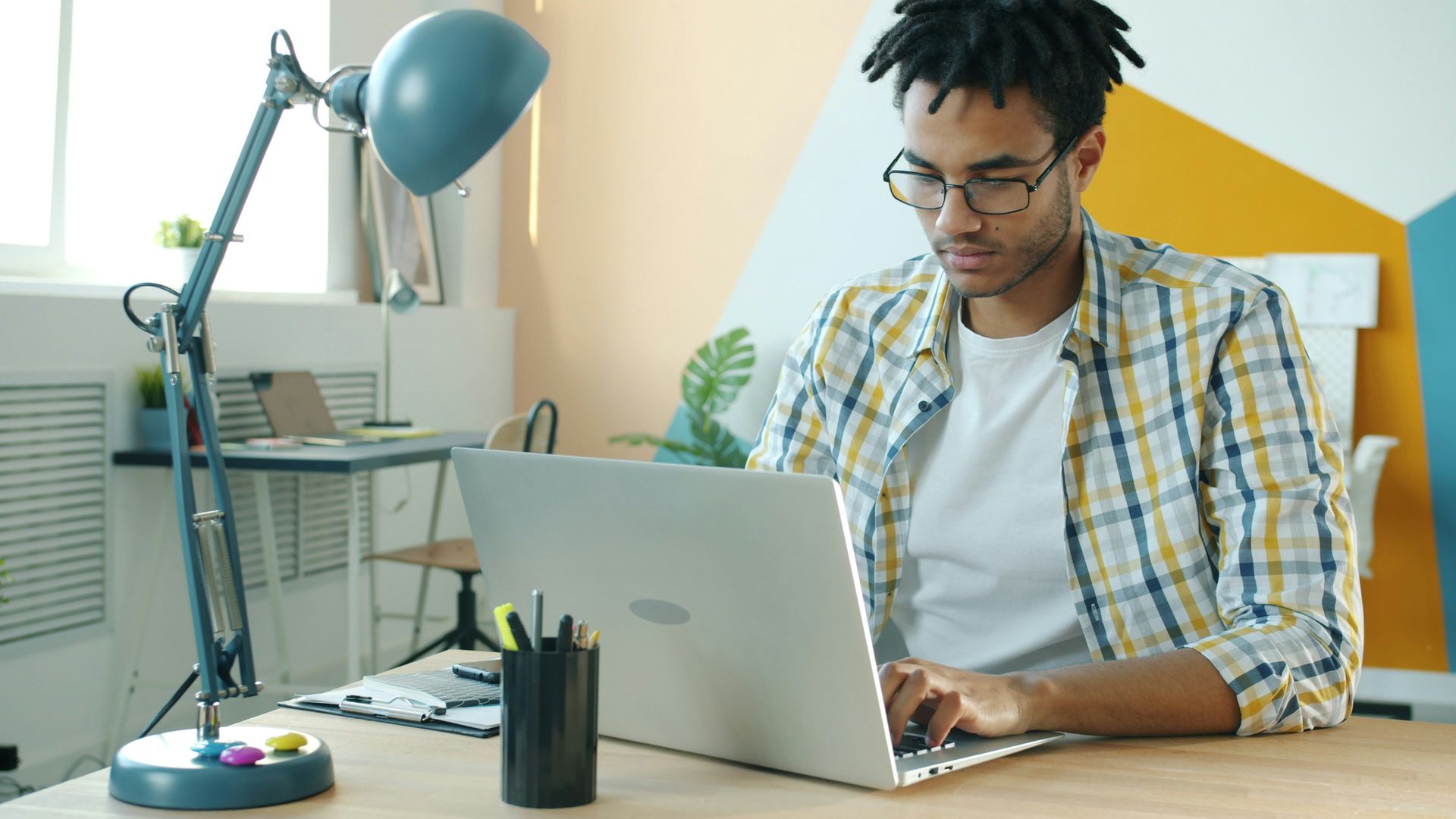 Man with glasses working on a laptop at a desk, yellow and blue decor.