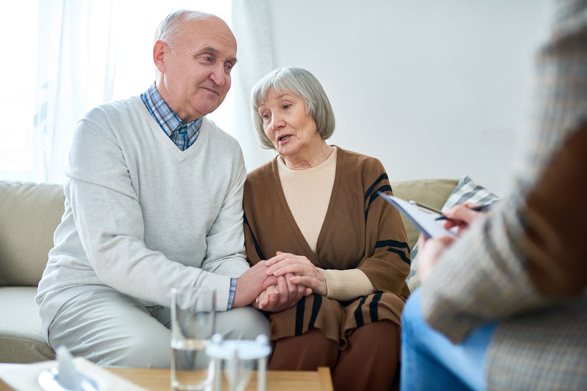 An elderly couple is sitting on a couch holding hands and talking to a doctor.