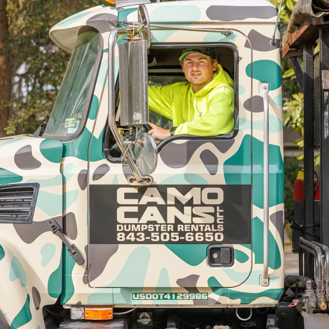 Man in neon yellow shirt drives a camo dumpster truck.
