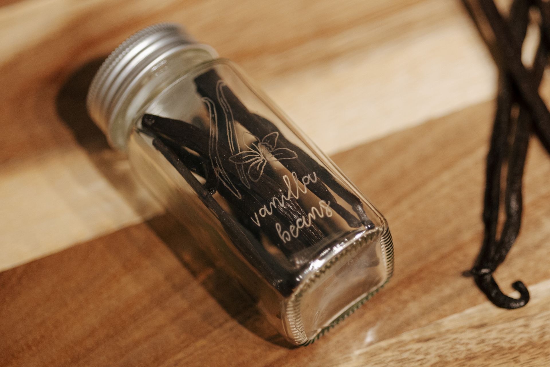 Glass jar of vanilla beans on a wooden surface; vanilla beans are scattered beside it.