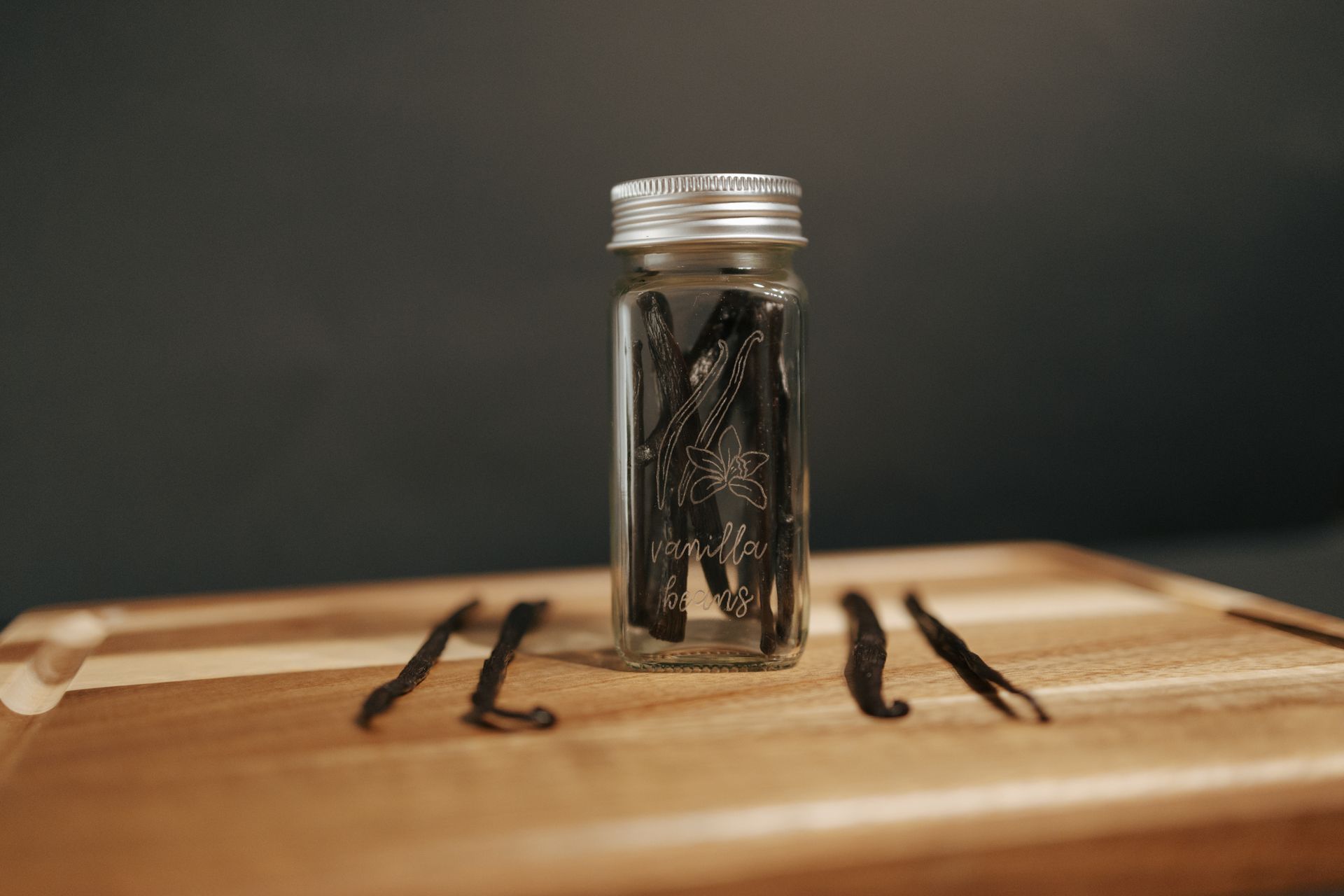 Vanilla beans in a glass jar on a wooden board, with a few loose beans scattered nearby.