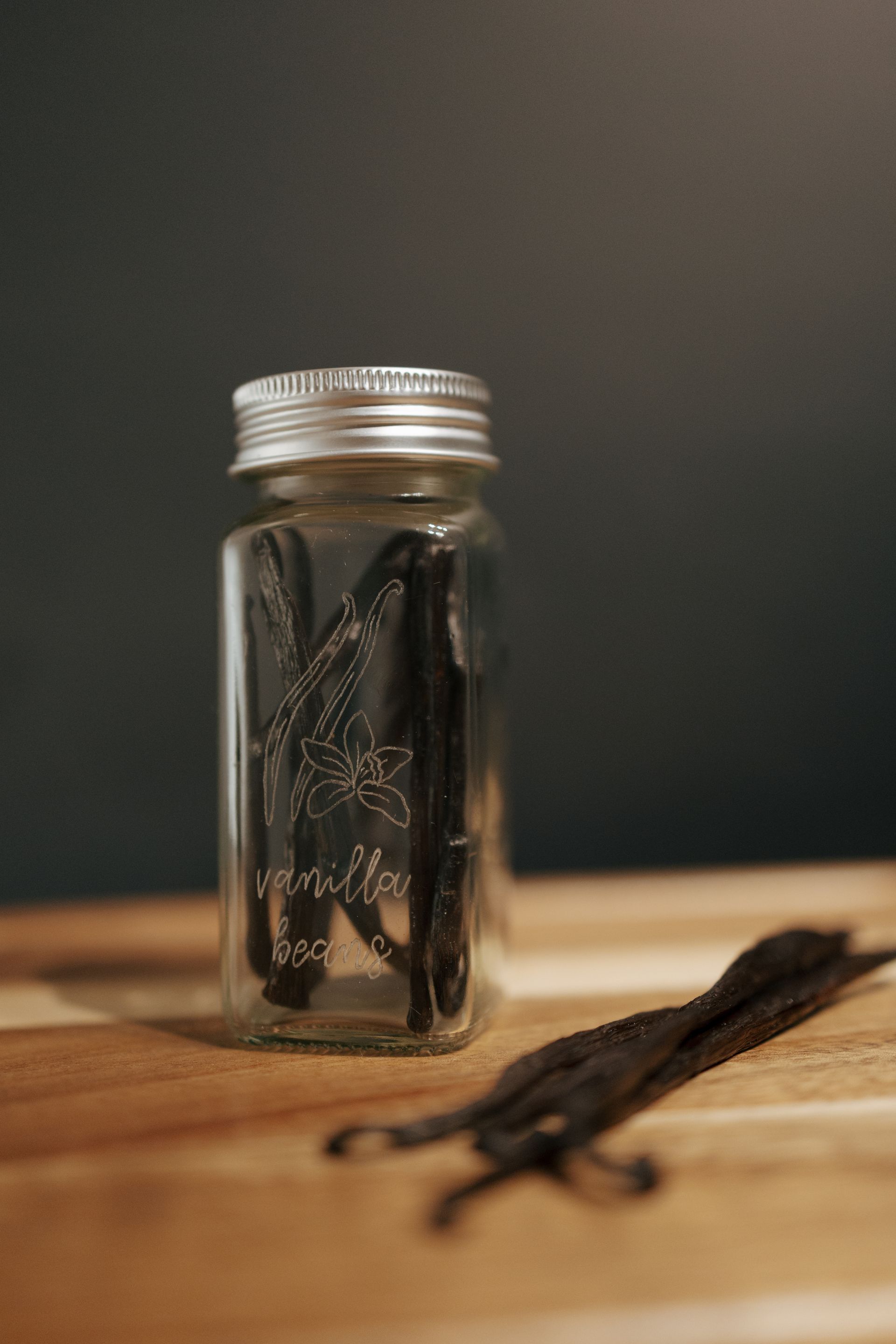 Jar of vanilla beans on a wooden surface, with several beans scattered nearby.