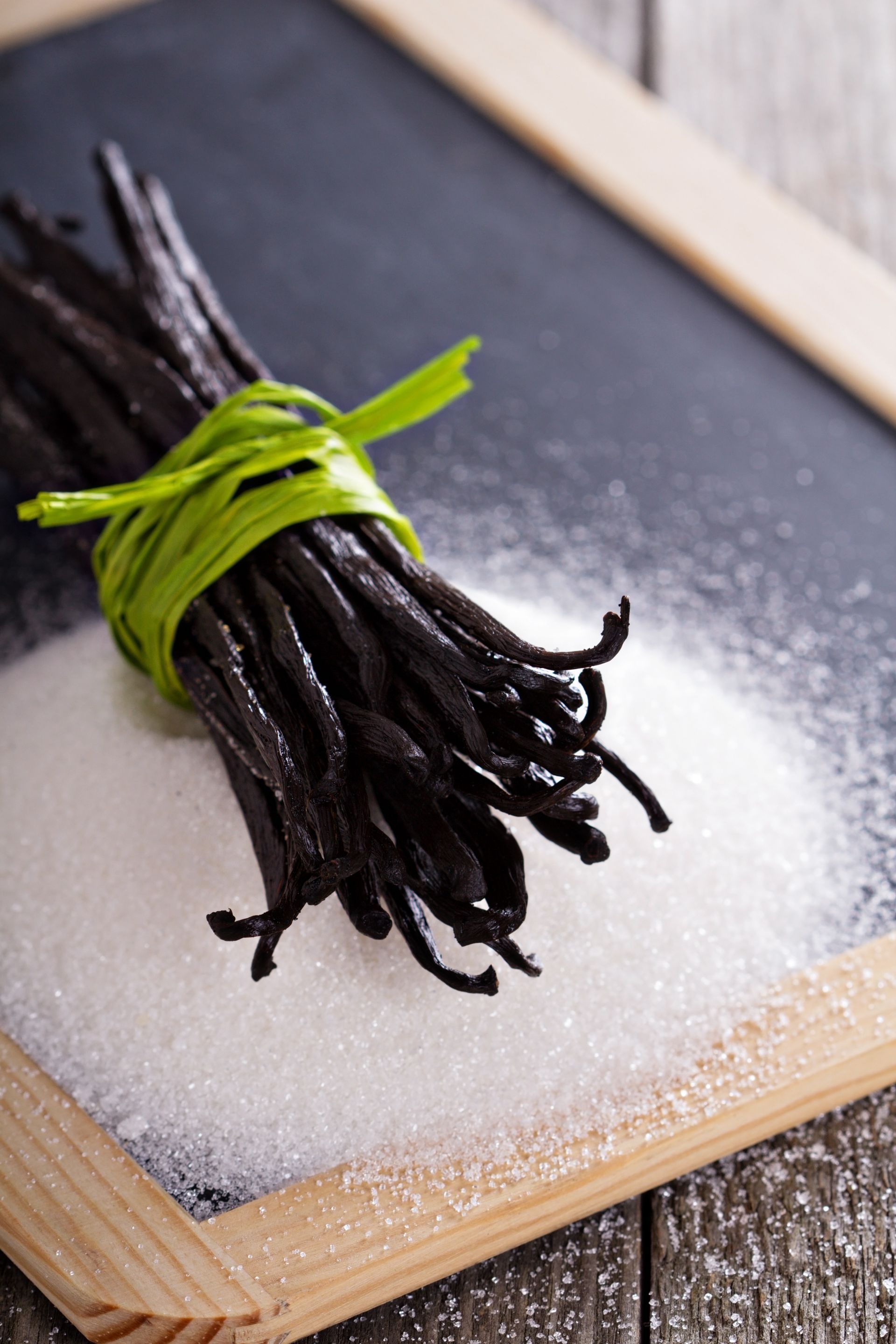 Vanilla beans tied with green grass on a pile of sugar, set against a black chalkboard.