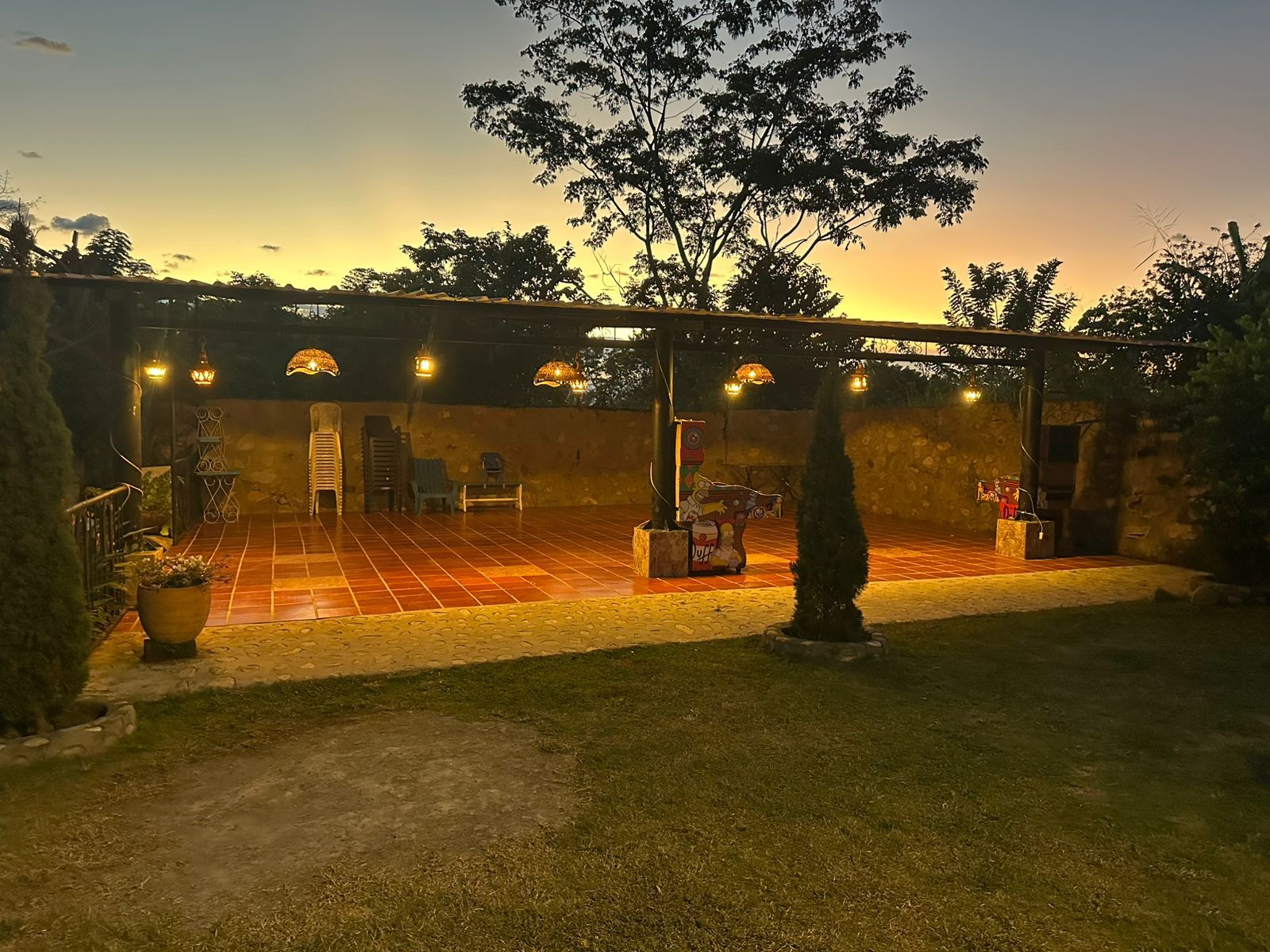 Patio al aire libre con luces de cadena, piso de ladrillo, grava y césped, iluminado por la puesta de sol.