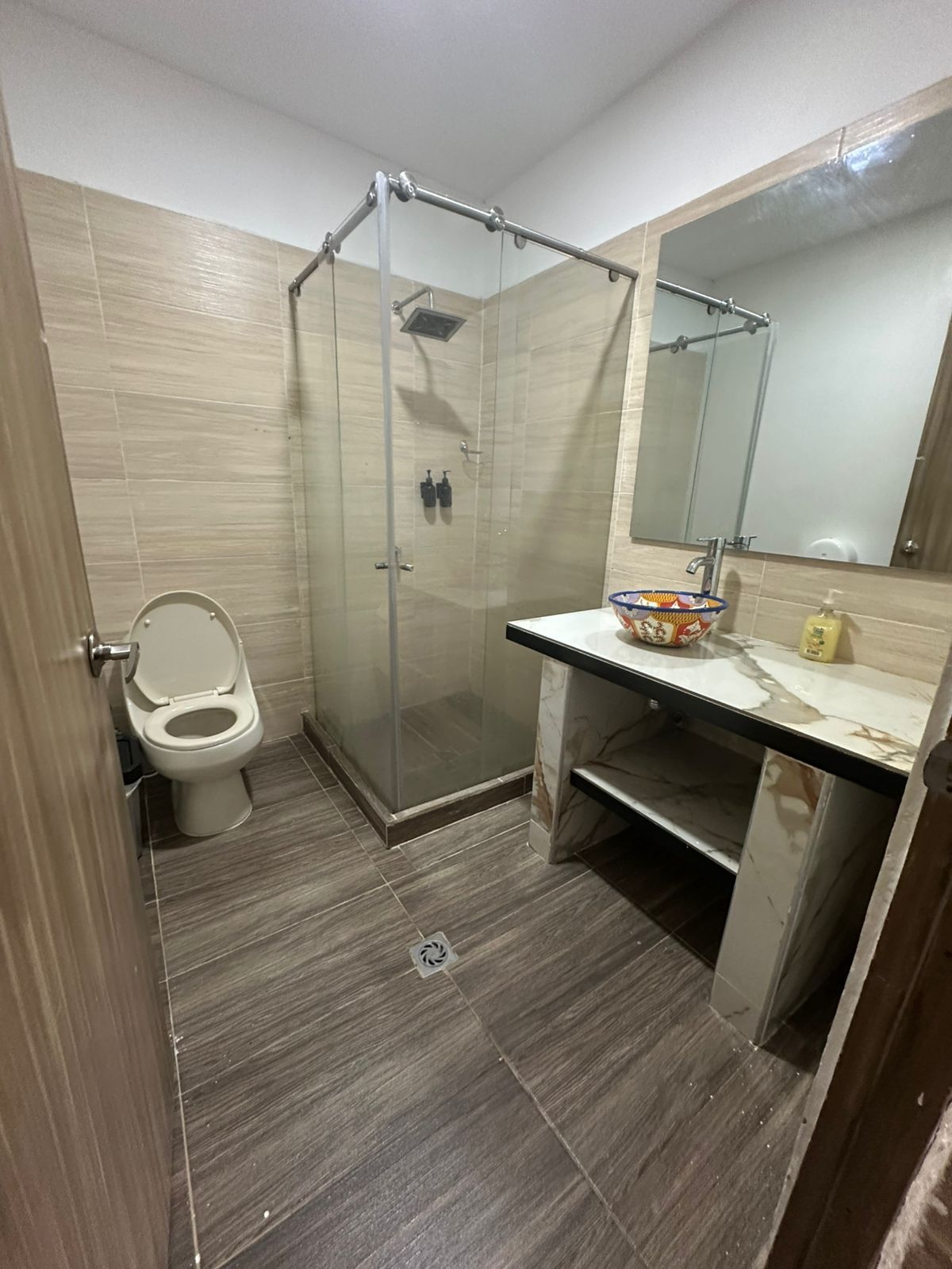 Baño con ducha, inodoro, lavabo y espejo. Azulejos en tonos tostados y marrones, y mampara de cristal.