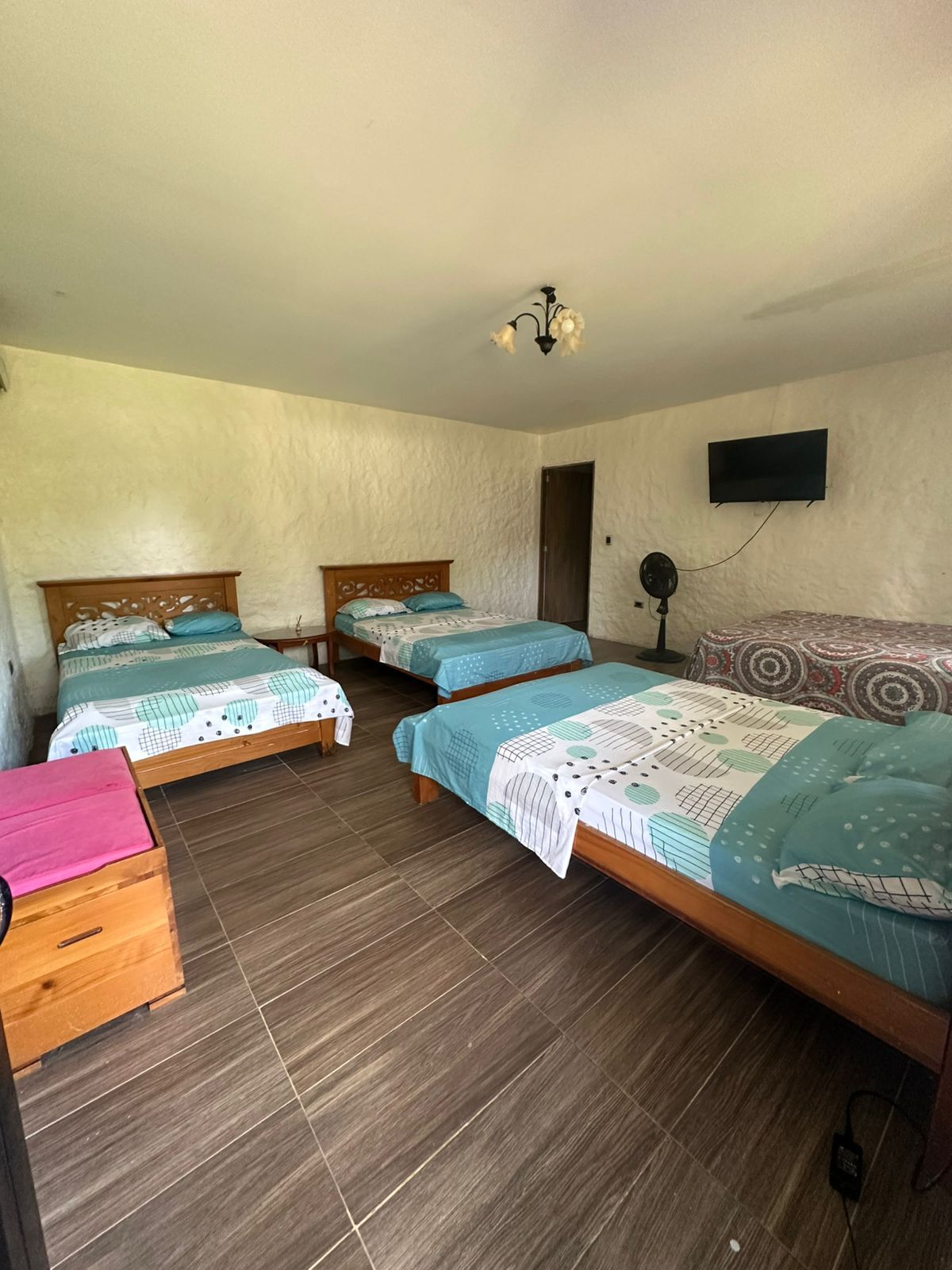 Dormitorio con cuatro camas; ropa de cama verde azulado y estampada. Baúles de madera y un ventilador a la vista.