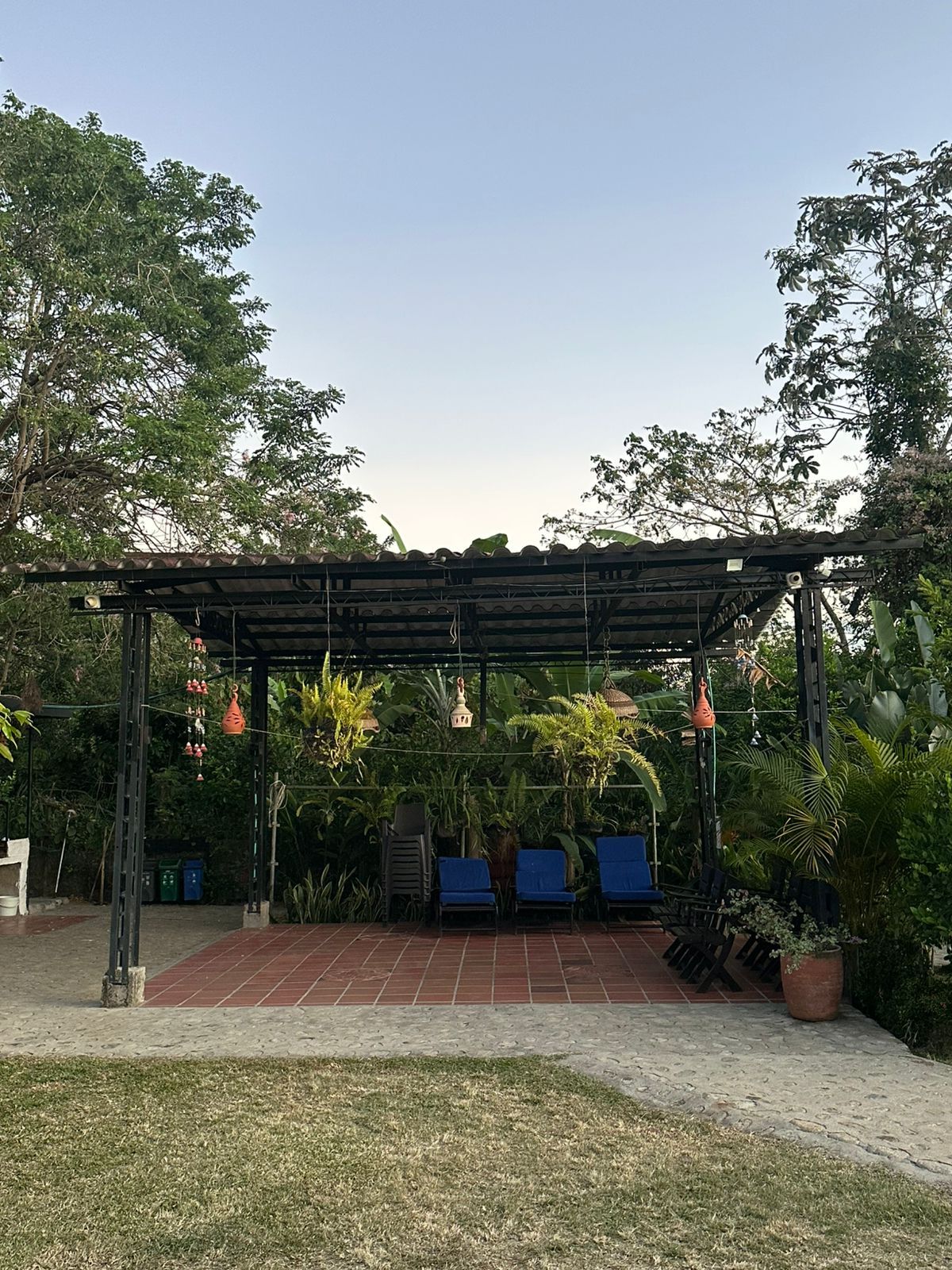 Patio con pérgola, piso de ladrillo, sillas azules y plantas colgantes; ubicado en un jardín con árboles bajo un cielo azul.