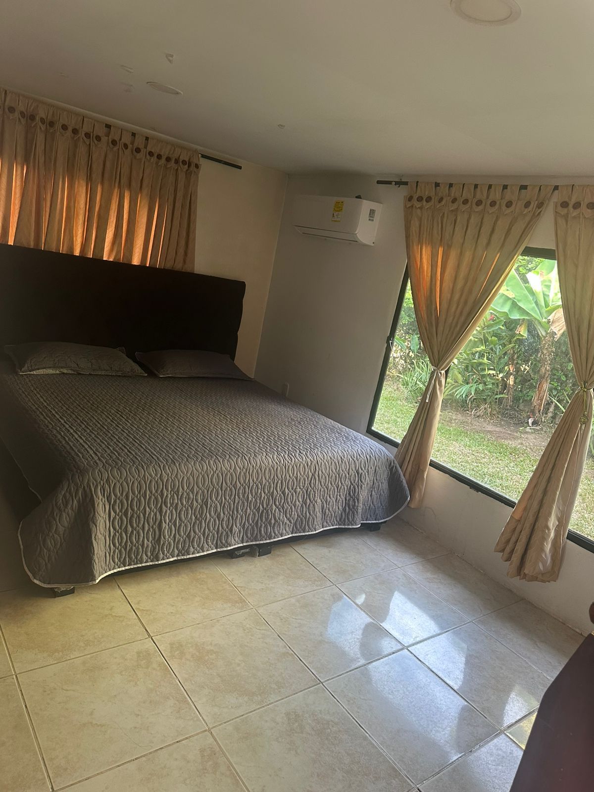 Dormitorio con cama, cortinas color canela, ventanal y suelo de baldosas.