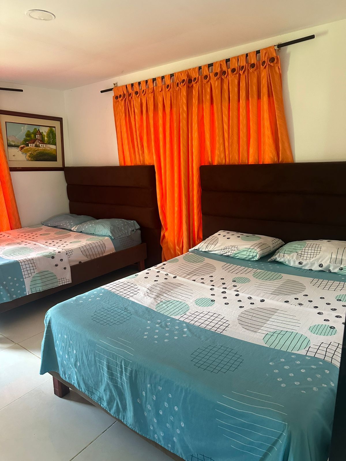 Dormitorio con dos camas, cortinas naranjas y un cuadro en la pared. Las camas tienen ropa de cama verde azulado y blanca.