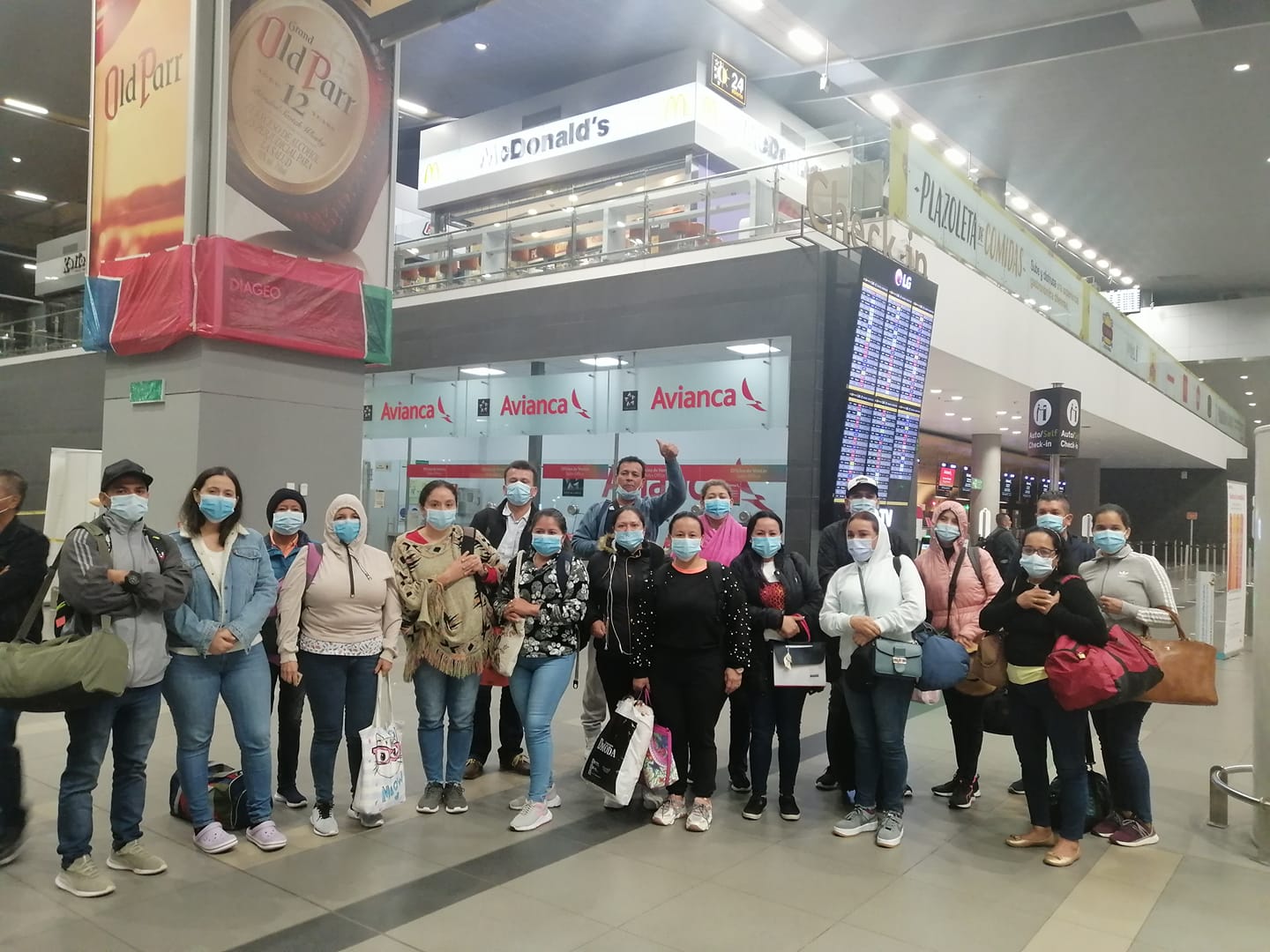 Grupo de personas con mascarillas en un aeropuerto. Algunas sonríen, saludan y llevan bolsas.
