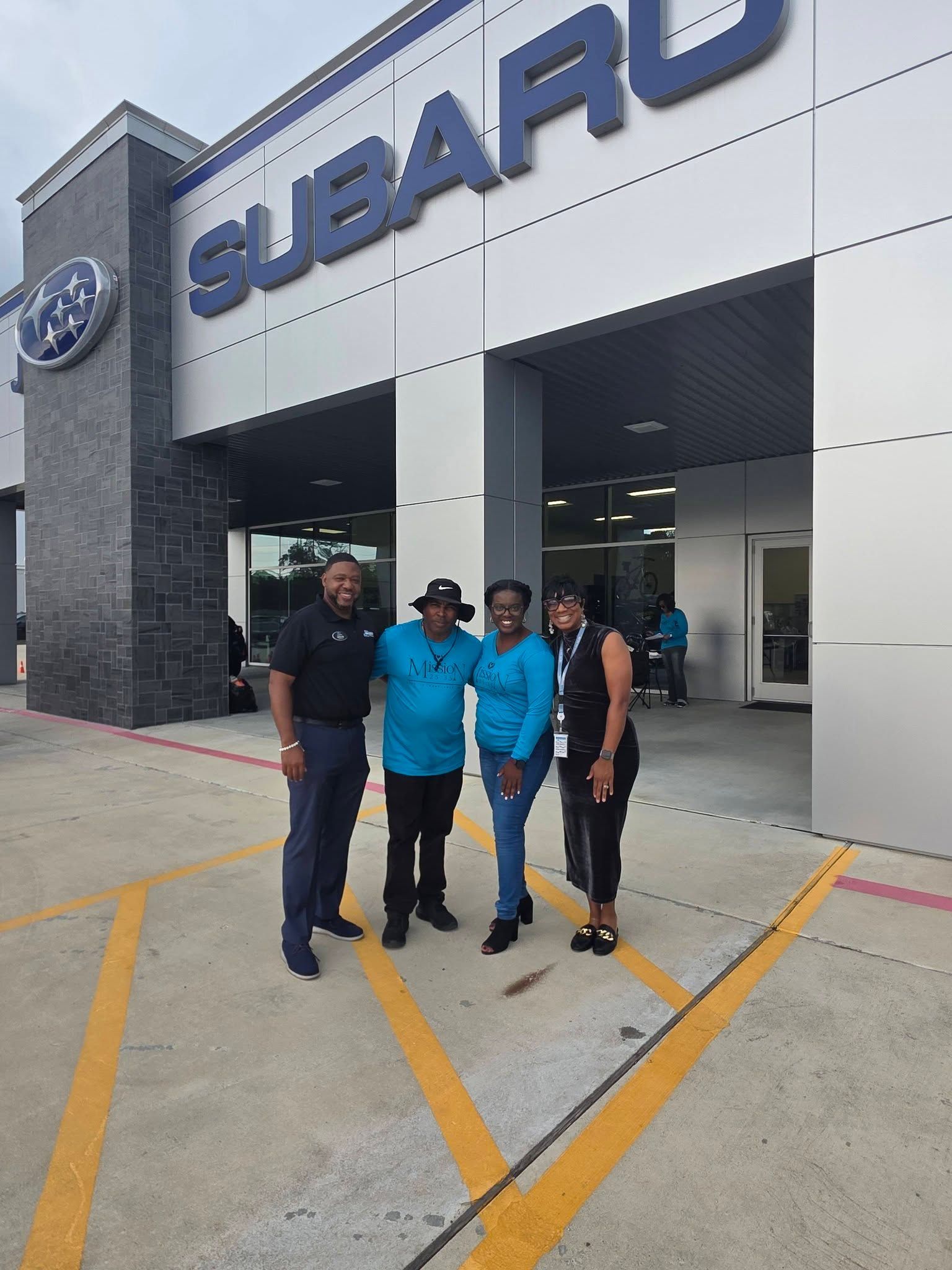 Four people stand in front of a Subaru dealership, smiling for the camera on a sunny day.