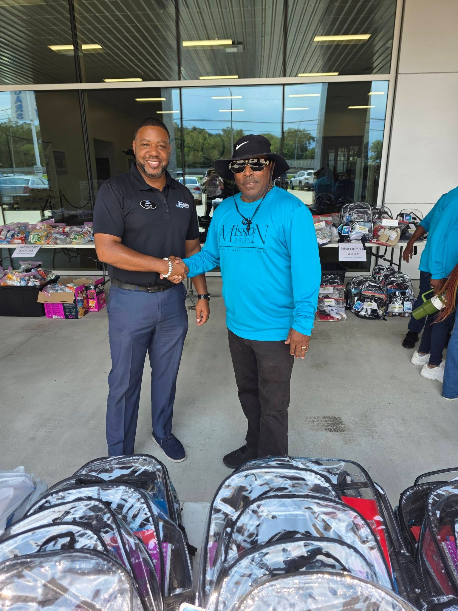 Two men shaking hands in front of tables filled with clear backpacks at an outdoor community event.