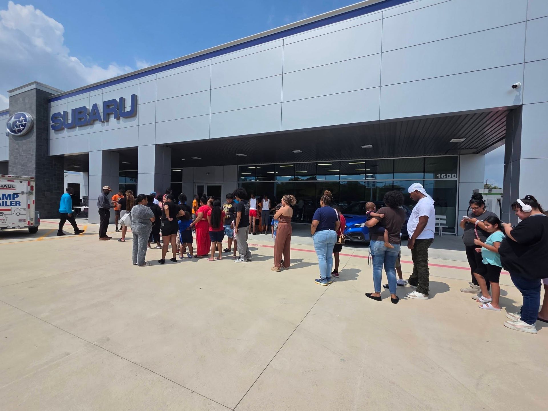 A line of people stands outside a Subaru car dealership on a sunny day.