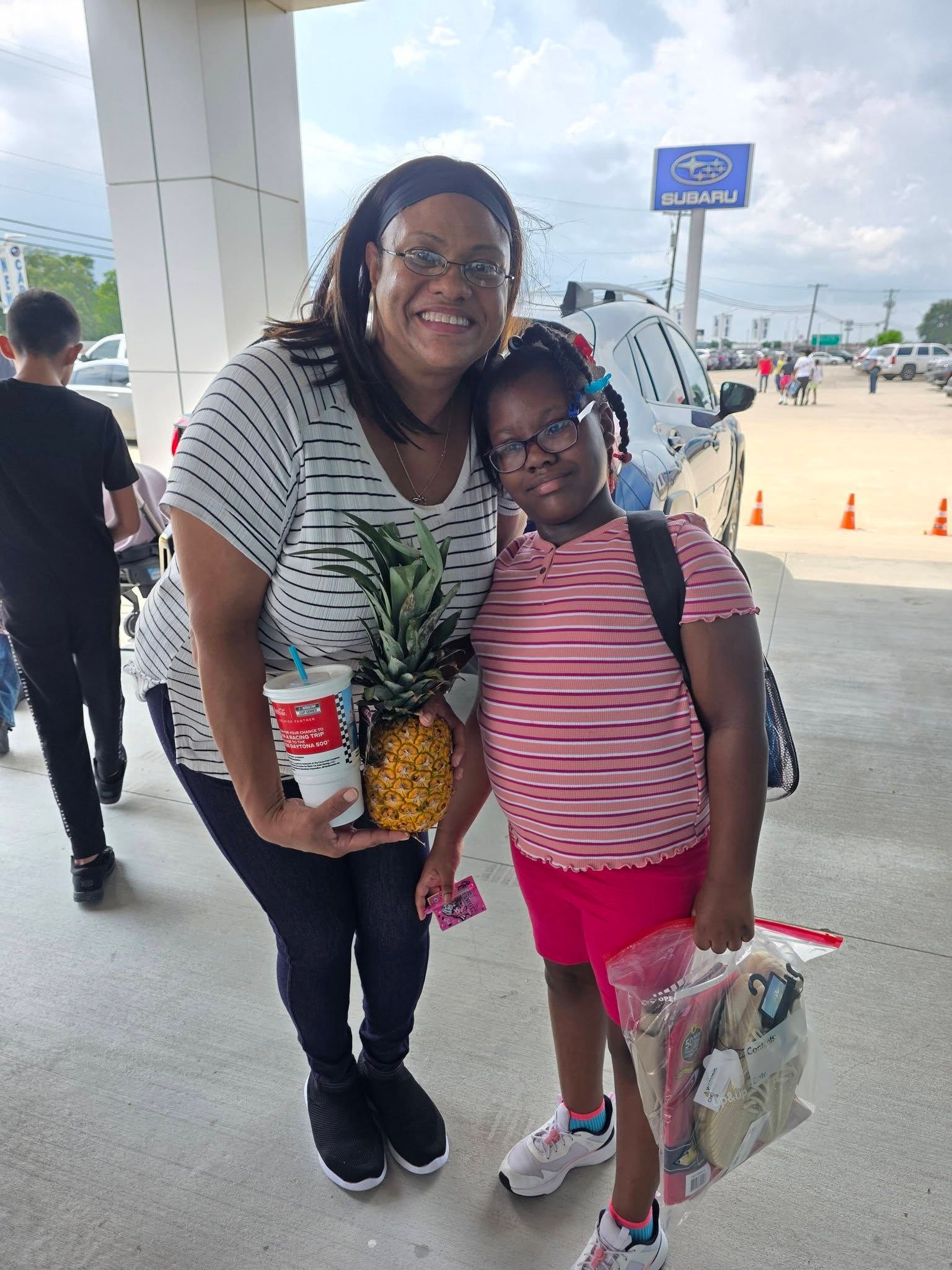 A smiling woman and child stand together outside near a parking lot, holding a pineapple and a plastic bag.