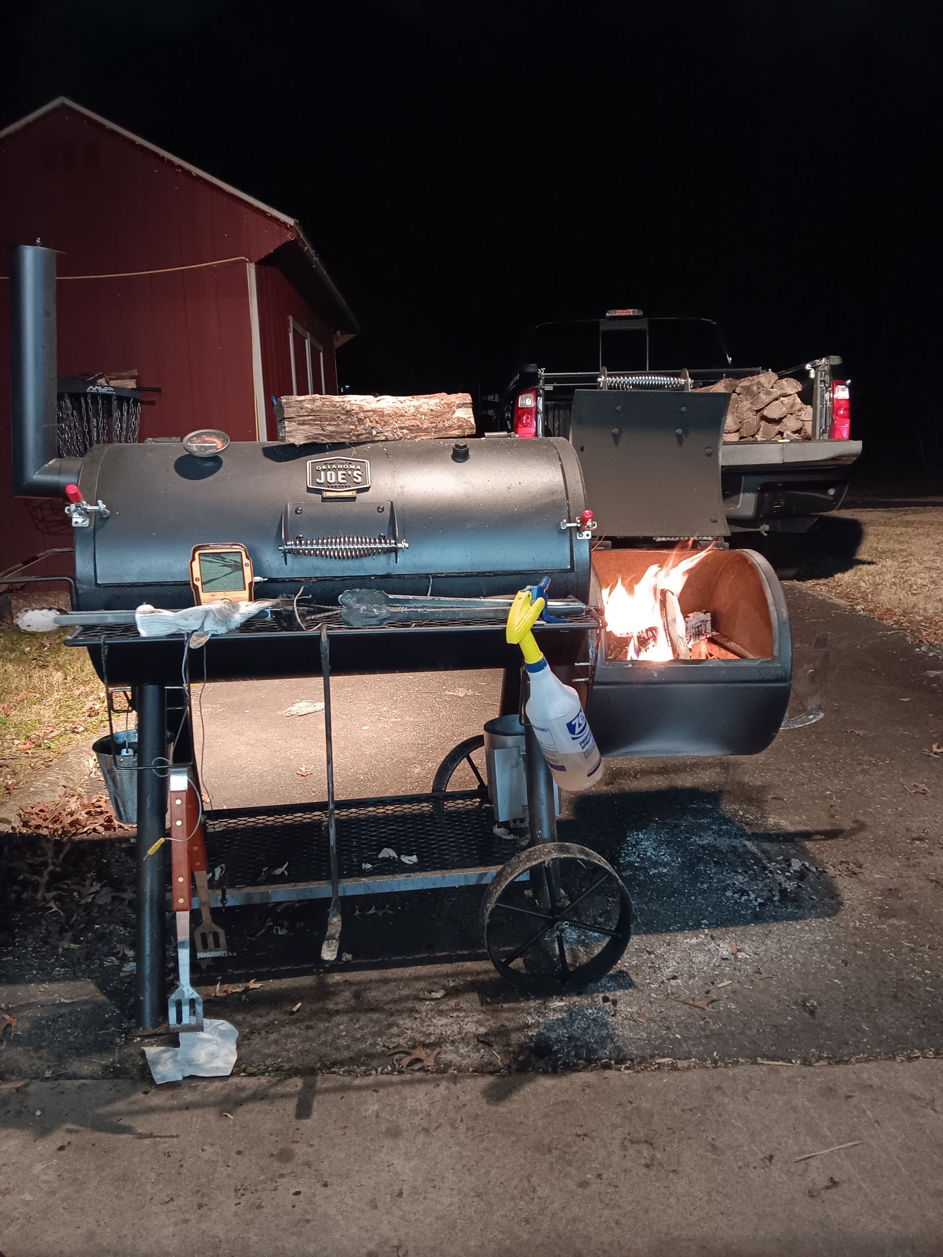 A smoker with fire glowing, next to a red barn at night with a pickup truck in the background.