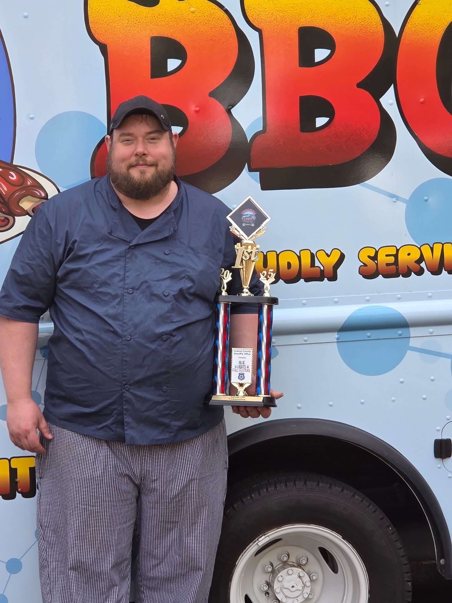 Man in chef attire holding trophy in front of BBQ food truck.