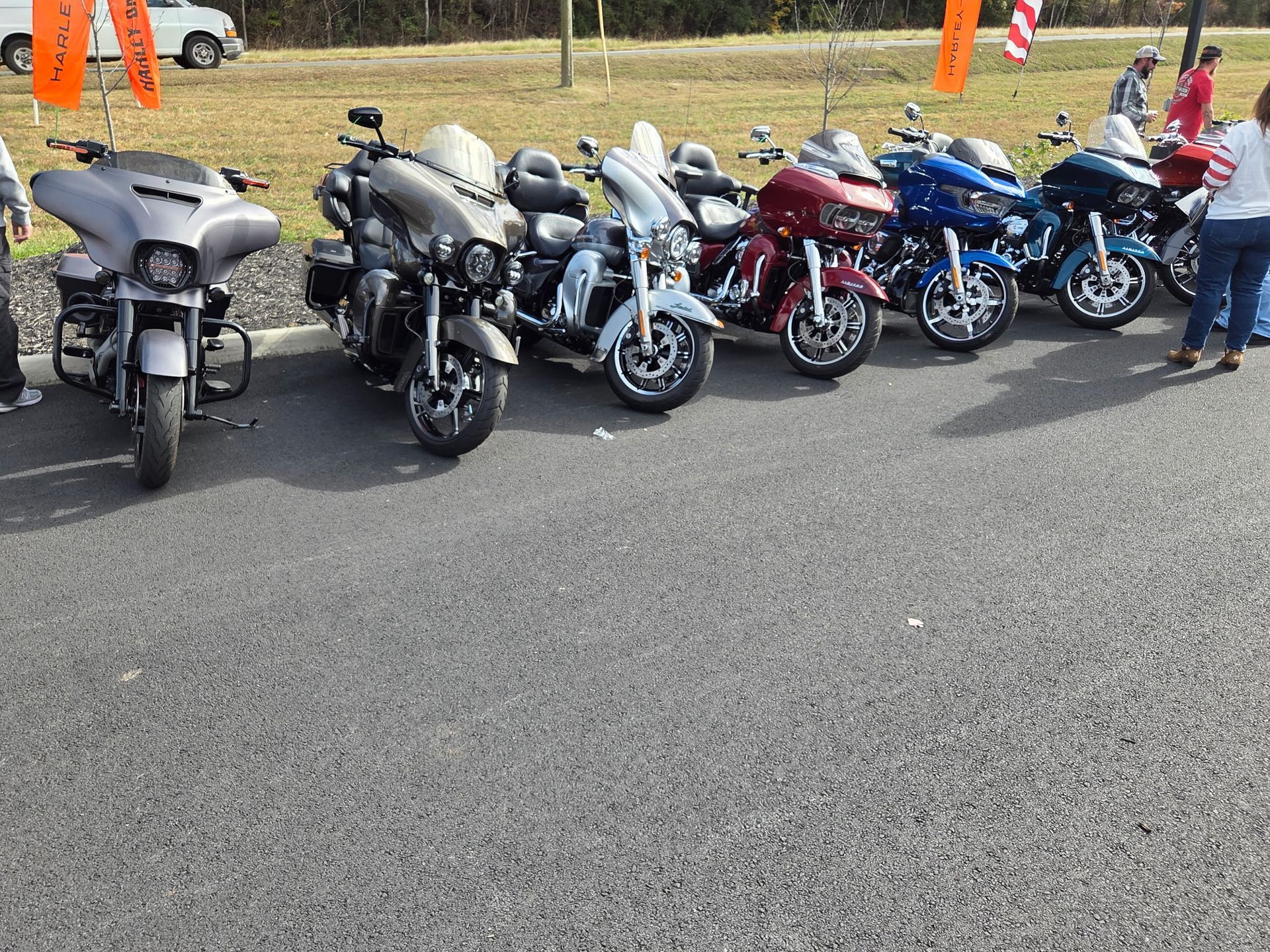Row of parked motorcycles in a paved area, various colors, with some people nearby.
